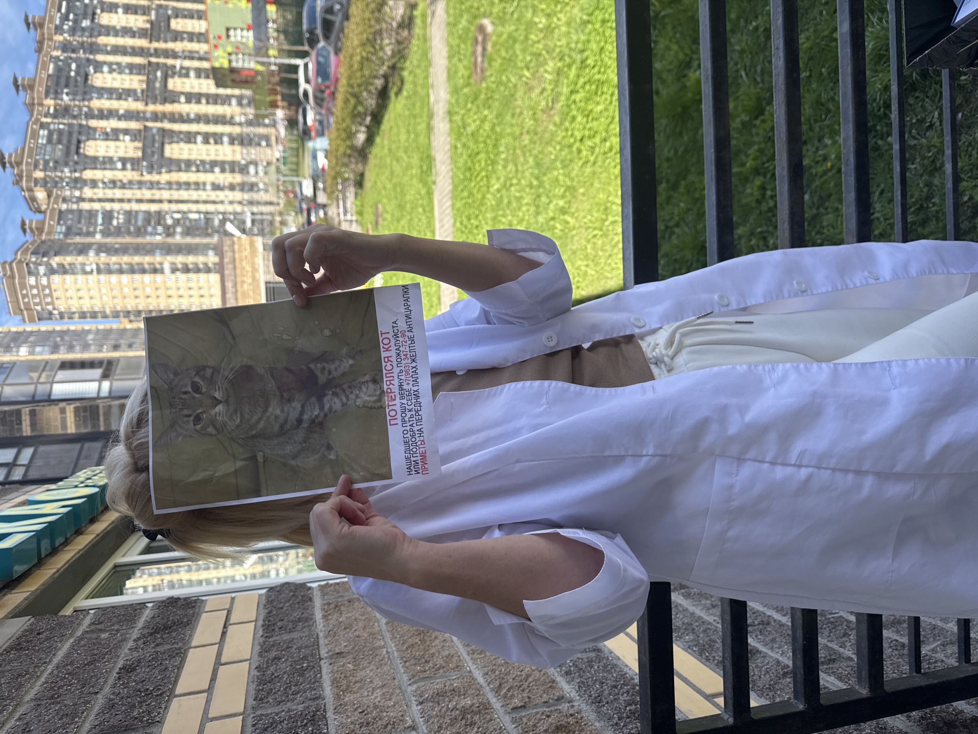 The image captures a person standing outdoors in Pargolovo, Russia, holding a "lost cat" poster that covers their face. The individual wears a white, long-sleeved garment, resembling a lab coat or oversized shirt, over a brown top and light-colored trousers. Their blonde hair is visible around the edges of the poster.
The poster displays a close-up photograph of a tabby cat and includes text in Russian. The visible text translates to: "LOST CAT. FINDER, PLEASE RETURN OR TAKE HOME +7(963) 347-72-90. FEATURES: YELLOW ANTI-SCRATCH CAPS ON FRONT PAWS."
The scene is an urban outdoor setting during bright daylight, indicated by the clear sky and visible sunlight. The person stands near a black metal railing, behind which lies a green grassy area and a street with several parked cars. In the background, tall, multi-story residential buildings with many windows dominate the skyline. The foreground features a brick wall and tiled pavement. The image conveys an activity of a person actively trying to find a lost pet in a residential area.