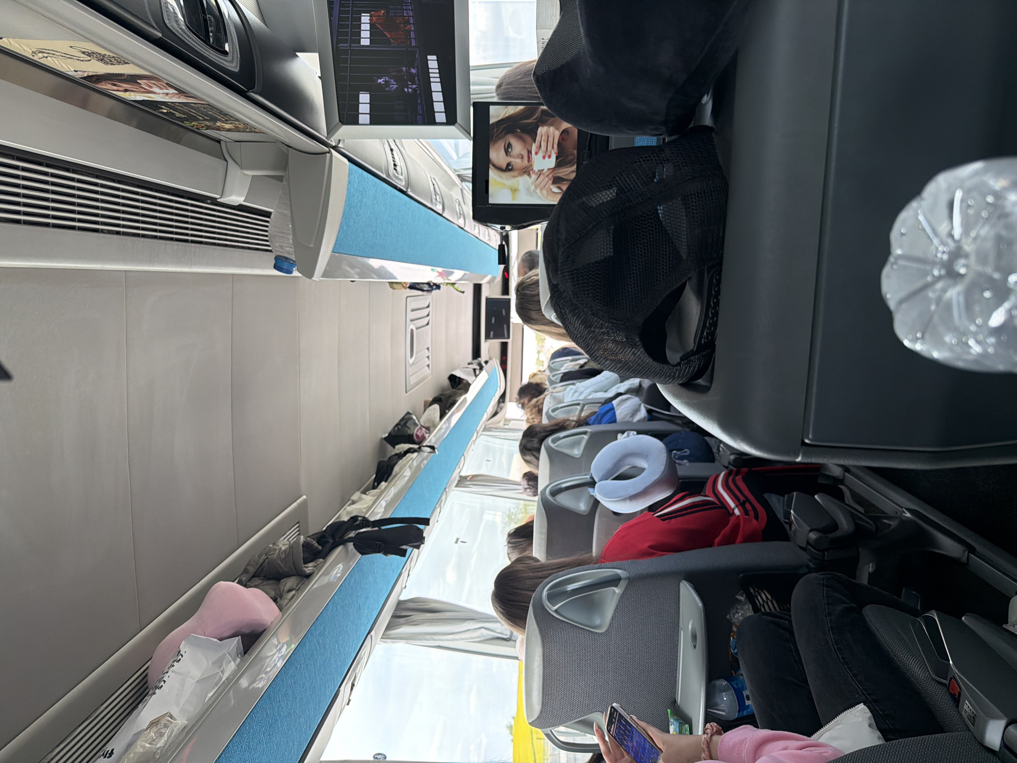 The image captures the interior of a modern passenger bus, likely traveling in or around Babaeski, Turkey, during daytime. The perspective is from an aisle seat looking forward and across the rows, with the image rotated 90 degrees counter-clockwise.
The bus features comfortable-looking grey seats equipped with individual entertainment screens. Several passengers are visible, mostly from behind or in partial views, seated and appearing to be engaged in travel. In the immediate foreground, a person is holding a smartphone, its screen showing colorful graphics. Another passenger further down the aisle is wearing a white neck pillow, indicating a readiness for rest or comfort during the journey.
Overhead storage bins, lined with a bright blue textured fabric, run along the ceiling. Various personal items are stored in these bins or on the seats, including a pink heart-shaped pillow, coats, and bags. A clear plastic water bottle is positioned near one of the screens at the front right.
Two individual screens are clearly visible: one prominently displays a woman holding a white cup, suggesting an advertisement or a scene from a movie/show. A second screen further down the aisle shows more abstract or game-like visuals. The bus interior appears clean and well-maintained. Bright natural light streams through the windows, indicating good weather outside. No discernible text is legible on any objects or screens within the image.