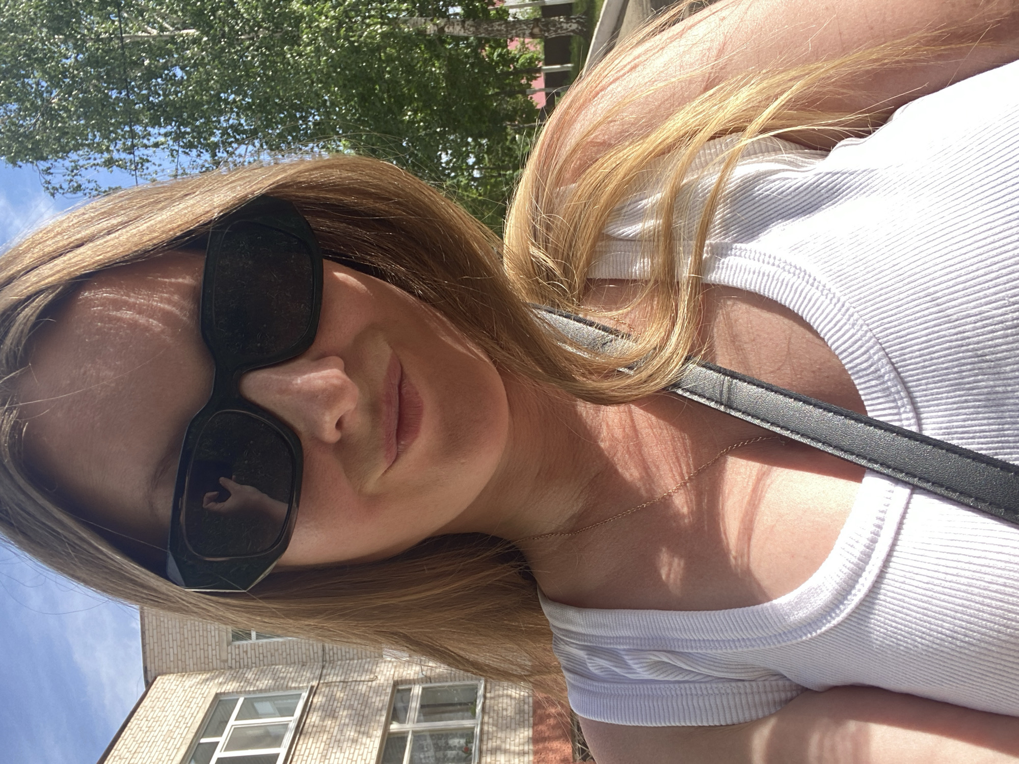 A selfie photo was taken of a woman with blonde hair wearing black sunglasses and a white ribbed tank top. A black strap is diagonally across her chest. The reflection in her sunglasses is a hand giving a hand gesture. The background is a building with windows, trees, and a blue sky. The photo appears to be taken outdoors, with sunlight creating shadows. The location is the city of Noviy, Russia.