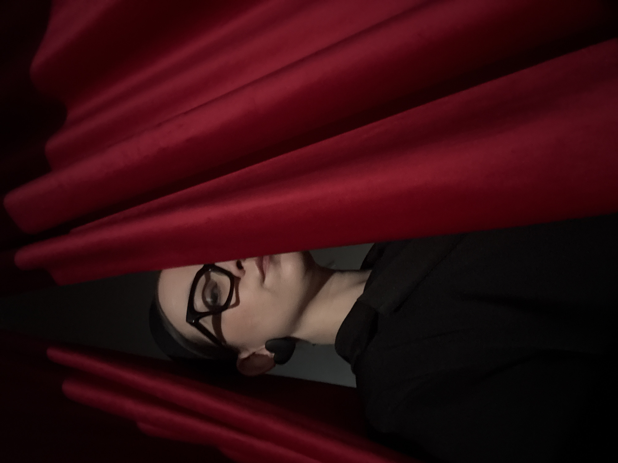 A person wearing glasses and a black outfit is peering through red curtains. They are oriented diagonally in the frame, with the curtains above and below them. The lighting is subdued, casting shadows across the scene. The person appears to have a black, heart-shaped object near their ear. No information about the location is detectable from the image.