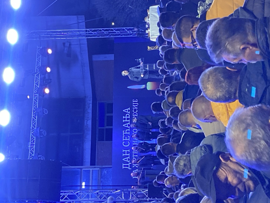 The image depicts an outdoor nighttime public gathering in Vranje, Serbia. A male speaker, dressed in a dark suit and glasses, stands at a clear podium equipped with a microphone and a glass of water, positioned on a raised stage. Behind him, a large blue backdrop displays the Cyrillic text "ДАН СЕЋАЊА НА ЖРТВЕ НАШЕ РЕПРЕСИЈЕ" (Day of Remembrance for the victims of our repression) and a graphic of a single lit candle with an orange flame. To the speaker's left, approximately six other men in dark attire are arranged on the stage. To the far right of the stage, a white-draped table holds various refreshments. Overhead, a metal truss system supports multiple stage lights, which cast a predominant blue illumination across the scene. A multi-story building with visible windows is partially discernible in the background, behind the stage structure. A dense crowd of adults, primarily facing the stage, occupies the foreground and midground, with attendees dressed in various jackets and coats.