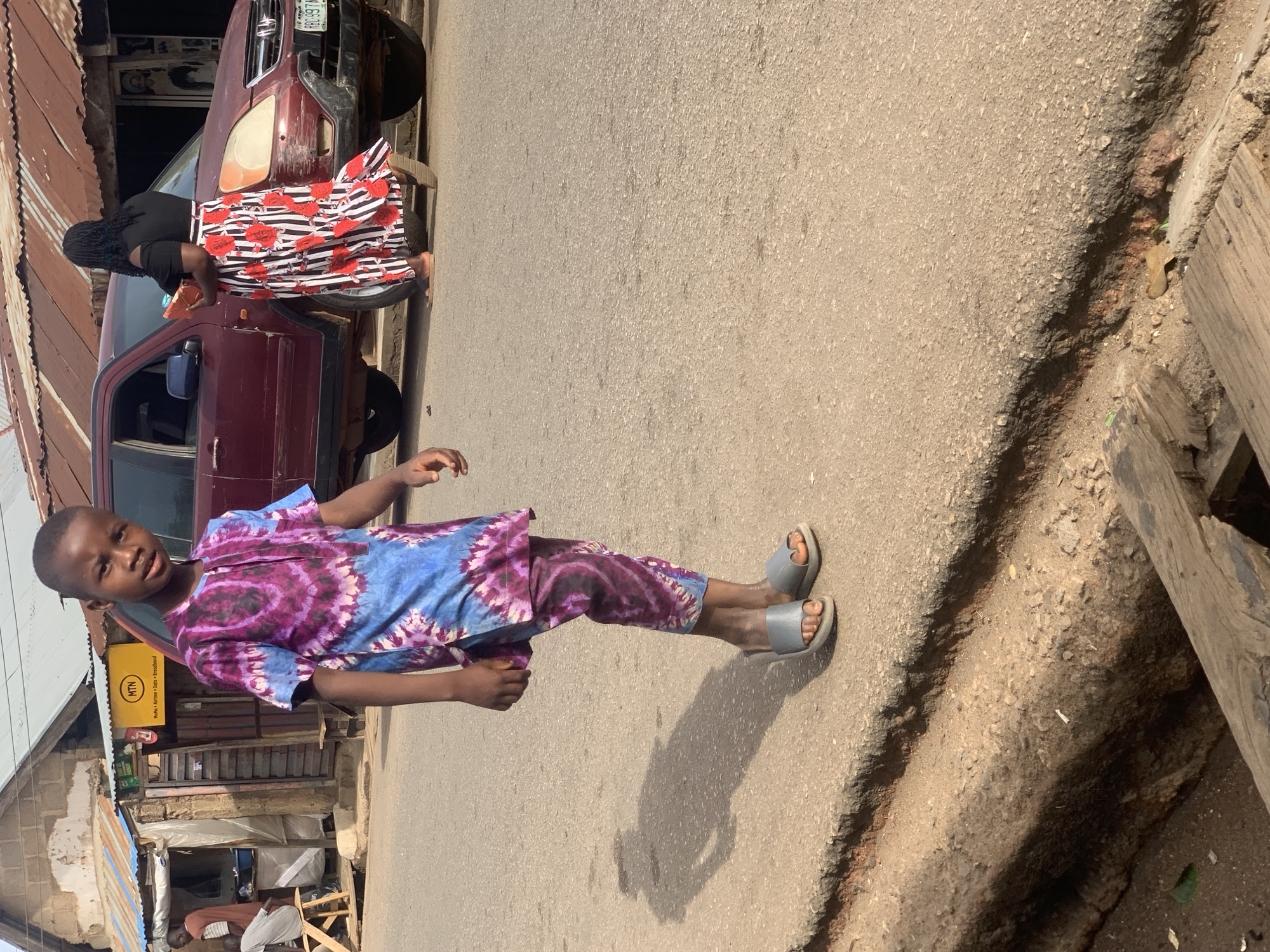 A young boy in a tie-dye purple and blue outfit and grey sandals stands on a paved road. In the background, a woman in a black top and a red, white, and black striped skirt leans over a maroon car. The car's license plate is partially visible. To the left are buildings with corrugated metal roofs. The boy is facing the camera with a neutral expression. An MTN sign is visible in the background. The location is likely in the city of Jos, Nigeria.