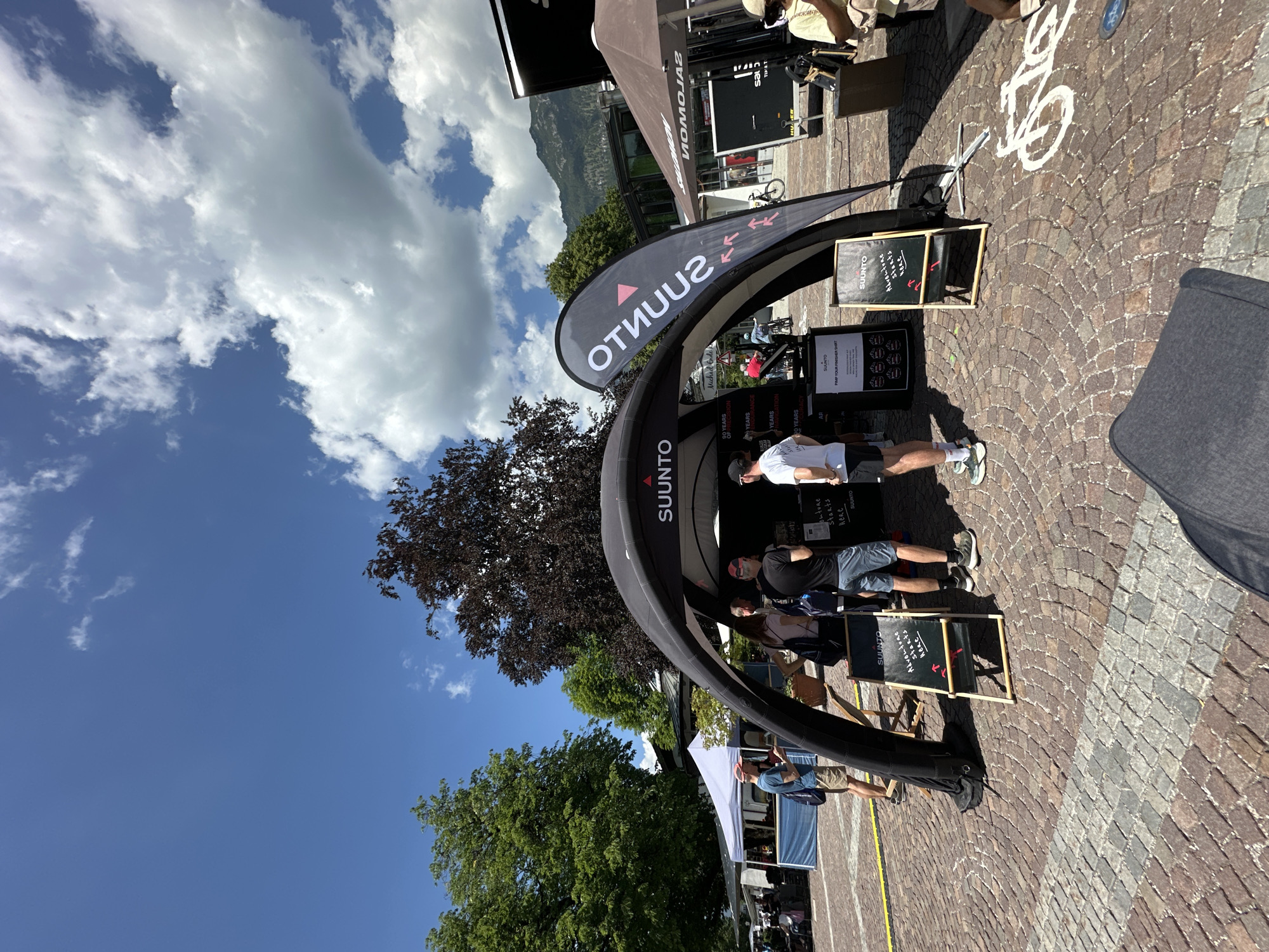 The image depicts an outdoor event, likely a market or festival, with a focus on the outdoor sports brand Suunto. A large, black inflatable archway tent with the Suunto logo is prominently displayed. Several people are gathered around the tent, some appearing to be employees or brand representatives interacting with visitors. There are two standing chalkboards with the Suunto logo and text. One chalkboard, to the left of the tent entrance, displays "SUUNTO" and then some bullet points. Another chalkboard in front of the tent reads "SUUNTO" and below it, "Adventure starts here" with two red arrows pointing downwards. Other elements visible include a white tent in the background, trees, buildings, and mountains in the distance, suggesting an alpine setting. The ground is paved with cobblestones. The sky is blue with scattered white clouds. The presence of mountains and the style of buildings suggests Garmisch-Partenkirchen, Germany.