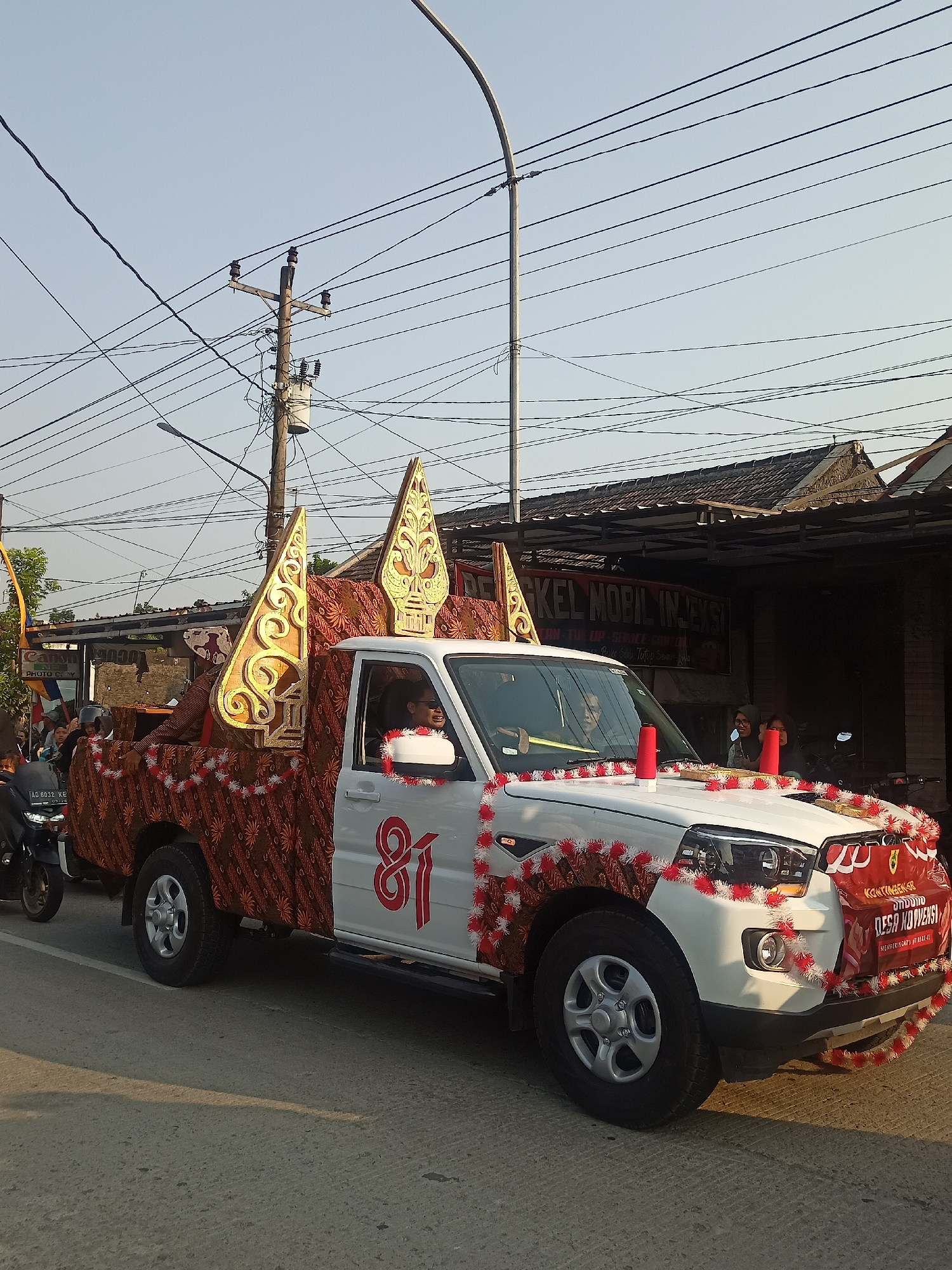 In the city of Ngrompongan, Indonesia, a white pickup truck, decorated with traditional Indonesian batik fabric and festive red and white garlands, participates in a parade. The truck is adorned with large, golden, ornate wooden headdresses, resembling those used in Wayang Kulit performances. A number "81" is prominently displayed in red on the side of the truck. A banner on the front of the vehicle reads "KONTINUETIKAR UNDANG DESA KOWENSI MERUPAKAN AKHLAK-A". The truck is moving on a paved road, with a clear sky overhead and utility poles with numerous wires lining the street. Buildings with tiled roofs are visible in the background. Several people are present in and around the truck, with some seated inside and others on the sides, seemingly part of the procession. A motorcycle is visible to the left of the truck. The scene appears to be a cultural or celebratory event.