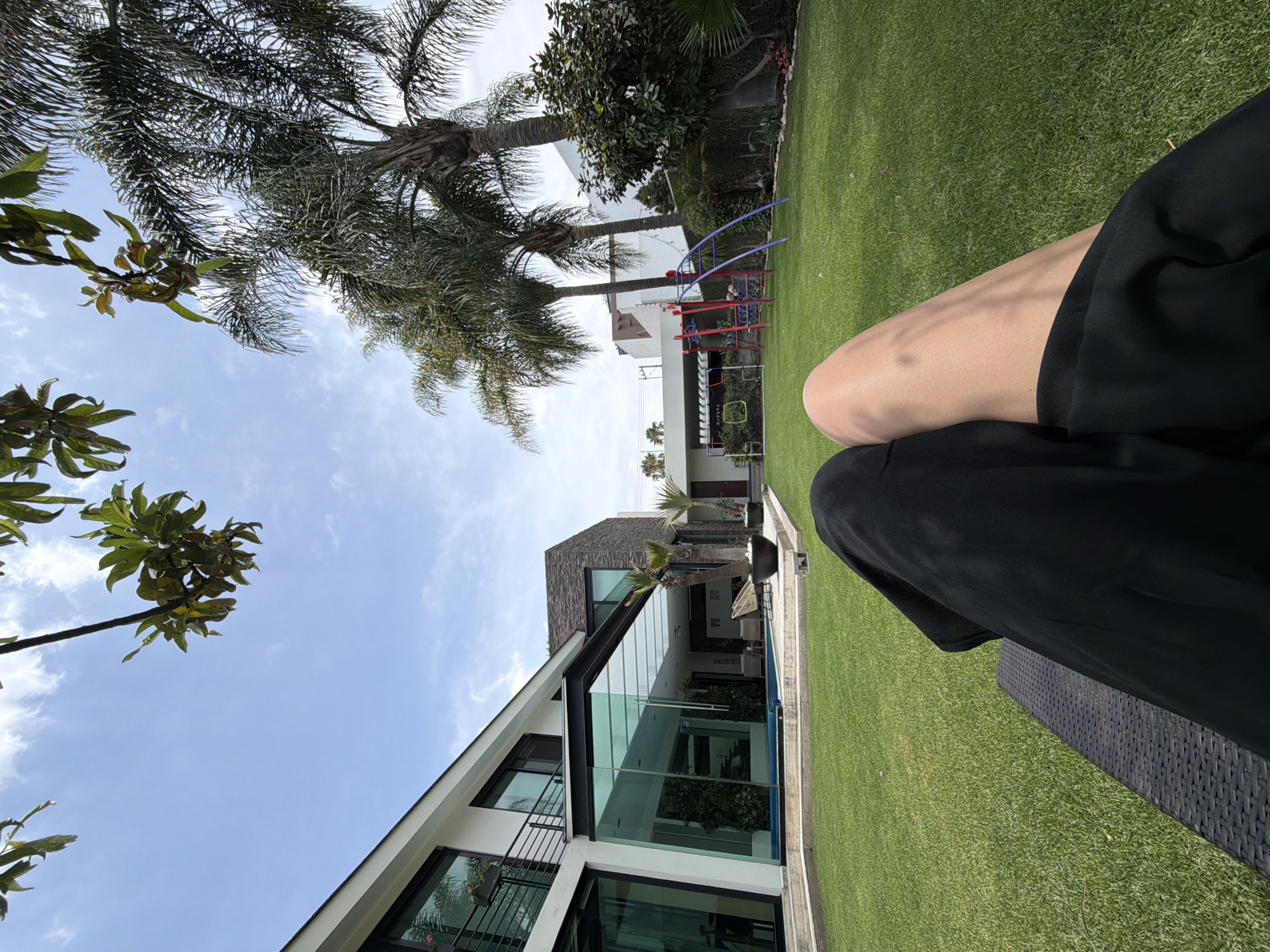 The image, taken outdoors in an unidentified city, showcases a modern house with large glass windows, a well-manicured lawn, and palm trees reaching towards a partly cloudy sky. Part of a person's leg and black clothing are visible in the foreground, suggesting the perspective is from someone seated. A playground structure can be seen in the background. The scene suggests a warm, outdoor setting on a bright day.