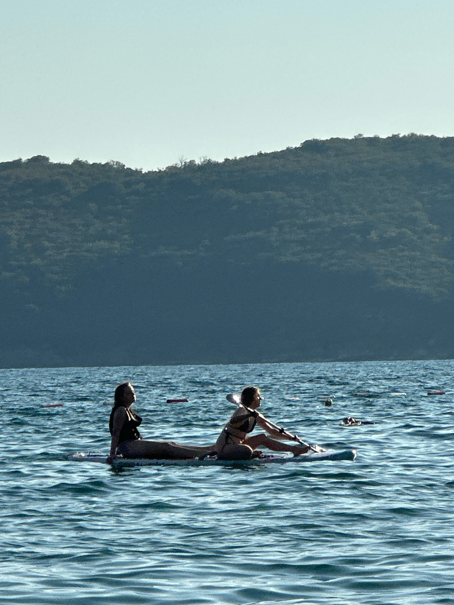 In the city of Boreti, Montenegro, two individuals are engaged in paddleboard yoga on the Adriatic Sea. The scene is characterized by calm, rippling blue water under a clear, pale blue sky. In the background, a dark, tree-covered mountain range slopes down towards the water's edge.
The two people are positioned on a single paddleboard. The person in the foreground, on the right, is kneeling and holding a paddle across their lap, appearing to be in a seated yoga pose. They are wearing a dark bikini. The person behind them, on the left, is also seated on the paddleboard, facing the same direction, and is wearing a black one-piece swimsuit. Their body is elongated along the length of the board, with their legs extended.
Several pink buoys are visible in the water, suggesting a designated swimming or recreational area. The overall atmosphere is serene and suggests a peaceful outdoor activity.
