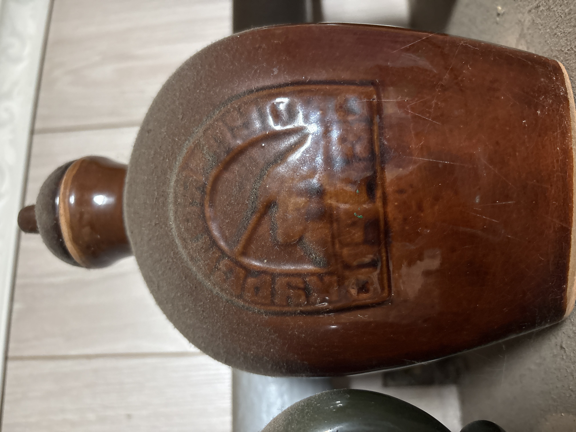 A close-up, eye-level shot shows a dusty, brown ceramic bottle with a curved body and a small cap. The bottle has an embossed circular design on its front featuring a silhouette of an animal, possibly a deer or a horse, within an archway. The embossed text around the design is partially obscured by dust but appears to read "ВЕТЛУЖСКИЙ" (Vetluzhskiy) at the top and "КРАЙ" (Kray) at the bottom. The background consists of light-colored vertical wooden planks, suggesting the bottle is on a shelf. A portion of another dark object is visible to the left of the bottle. The overall scene suggests an indoor setting, likely a storage area or collection.
