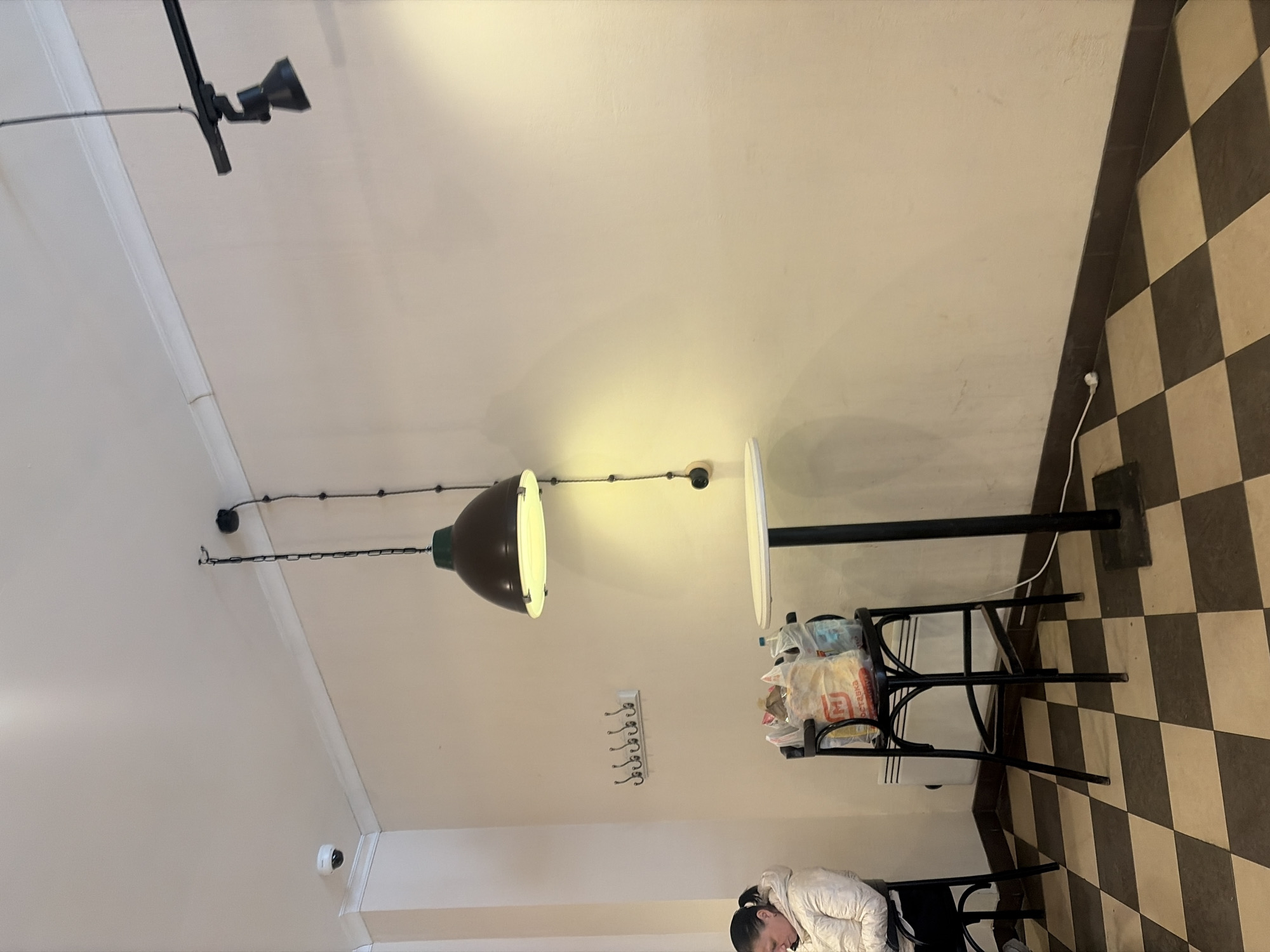 The image depicts an interior view, likely a cafe or restaurant, situated in Saint Petersburg, Russia, as evidenced by the checkerboard tile pattern common in older establishments.
The scene is primarily illuminated by artificial lighting, including a prominent pendant lamp with a dark shade hanging centrally in the frame and additional track lighting near the top of the image. The walls are light-colored, and there is a row of coat hooks mounted on one of them.
A small, round table with a black metal frame stands near the right side of the frame. It is topped with packages of food items.
At the bottom of the image, partially obscured by the table, is a person wearing a white puffer jacket. The individual appears to be seated or kneeling.
The floor features a distinctive checkerboard tile pattern.