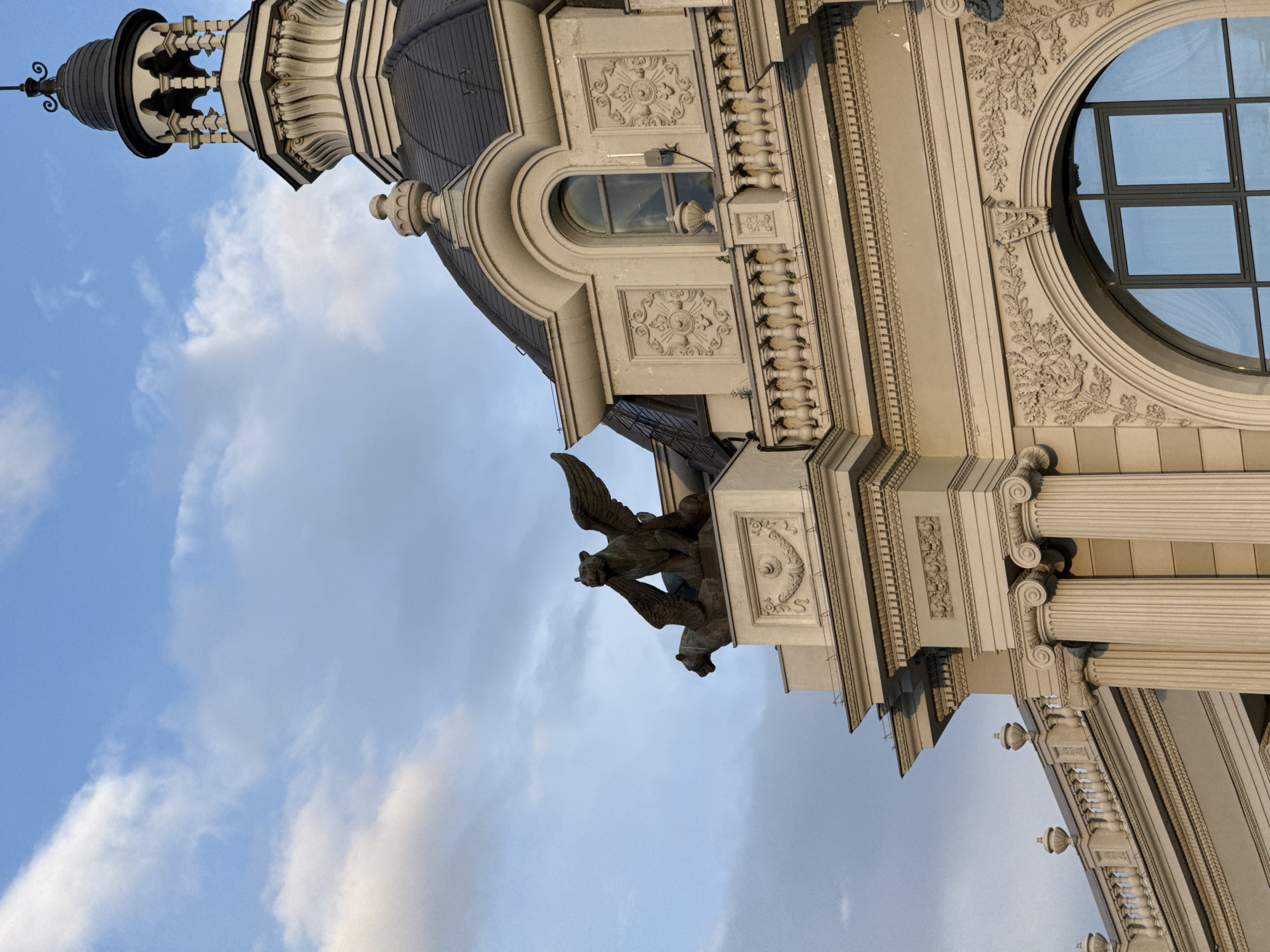 A low-angle shot of the Palace of Farmers in Kazan, Russia, angled from the lower left corner towards the top right corner. A blue and white sky is visible in the background. The building's facade is adorned with elaborate ornamentation, including sculpted eagles or gryphons with outstretched wings, decorative floral and geometric patterns, and neoclassical columns. The dome and arched windows are visible.