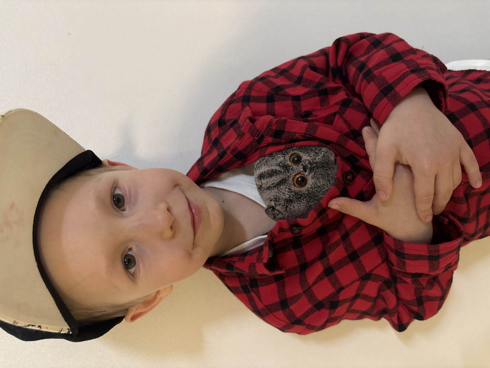 A young child is depicted in a medium close-up, gazing directly at the camera with a gentle smile. The child has light-colored hair, peeking out from under a beige baseball cap with a dark brim, worn backward. He is dressed in a red and black plaid flannel shirt over a white undershirt. Tucked into the chest area of his shirt is a grey plush toy, resembling a cat or owl, with prominent round, orange-yellow eyes, peeking out. The child's hands are clasped over the plush toy, holding it securely. The background is a plain, light-colored surface, possibly a wall or floor, indicating an indoor setting. The lighting is bright and uniform, illuminating the child's face and the details of his clothing and the toy. The image conveys a sense of warmth and playfulness, capturing a sweet moment. No text is visible in the image. This scene takes place in Chaikovsky, Russia.
