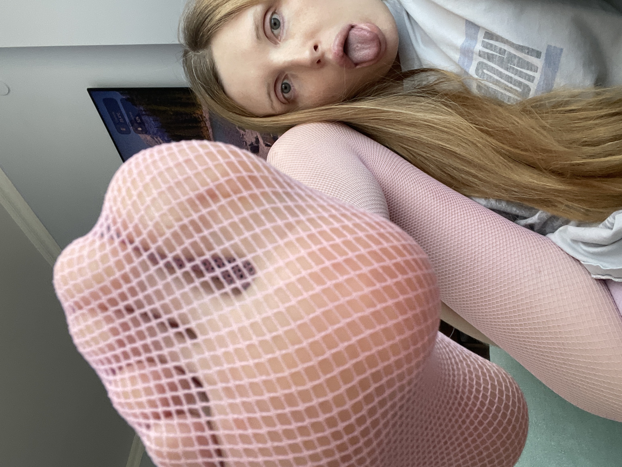 A young woman with long blonde hair is lying down, with her face close to the camera. She is sticking her tongue out and looking at the camera with wide eyes. Her head is tilted to the side. She is wearing a grey t-shirt with some text that is partially visible, reading "NYON". She is also wearing pink fishnet tights. A television screen is visible in the background, displaying a landscape image. The scene appears to be indoors, possibly in a home, with a neutral background. The lighting suggests daytime.