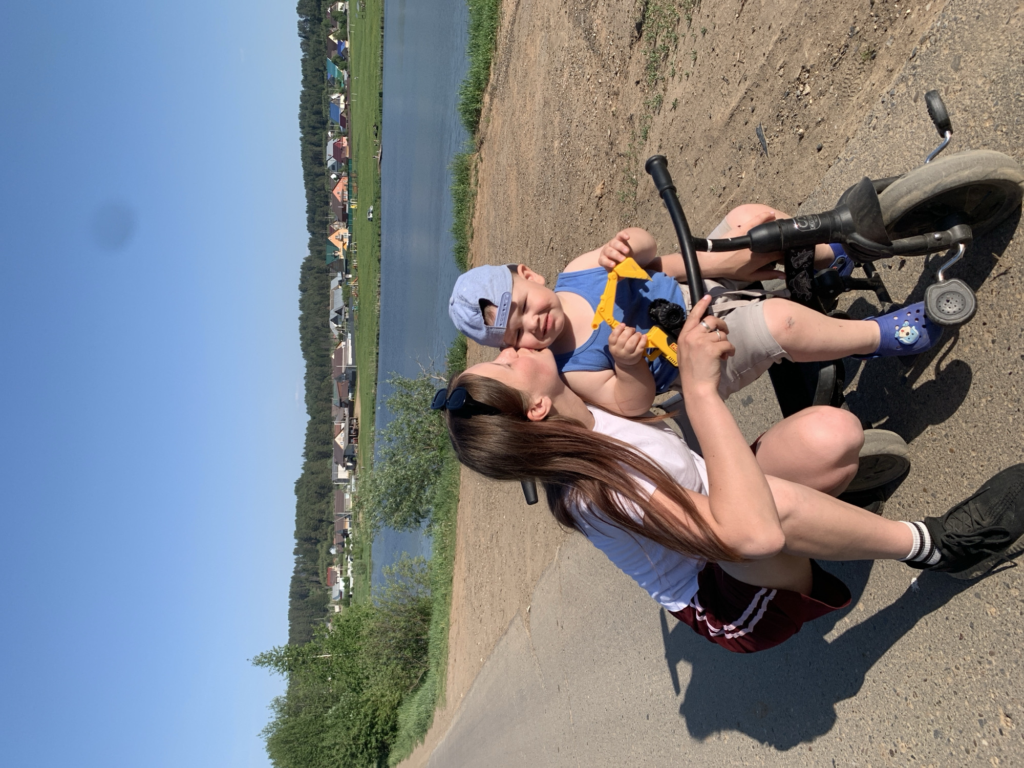 A woman and a young child are seated on the ground next to a tricycle. The woman is wearing a white t-shirt, maroon skirt, and black sneakers with white stripes on the socks. She has long brown hair and sunglasses perched on her head. She is kissing the child on the cheek. The child is wearing a blue tank top and cap, beige shorts, and blue Crocs. He is holding a yellow and black toy excavator. The tricycle is black and blue.
The background shows a landscape with a body of water, trees, and buildings. In the distance are houses with colorful roofs, which are consistent with the architectural style found in the city of Фоки, Russia. The sky is clear blue.