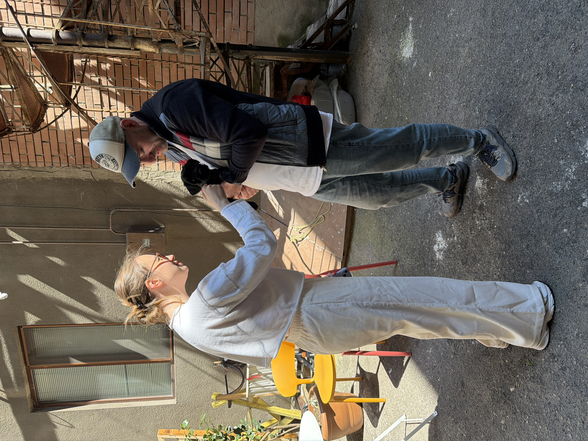 The image, taken in Tbilisi, Georgia, captures two individuals interacting in a sunlit urban courtyard or alleyway. The scene shows them standing on a concrete or asphalt surface, with strong shadows indicating bright daylight on a clear day.
The man, positioned on the left side of the frame, wears a grey and white baseball cap with a circular logo (text illegible), a dark zip-up jacket over a white t-shirt, and dark jeans with dark athletic shoes. He is looking down intently at a small, dark object held by the woman, his expression appearing engaged and possibly smiling.
The woman, on the right, has light brown hair tied in a bun and wears sunglasses on her head. She is dressed in a light-colored long-sleeved top and matching light-colored corduroy pants with light sneakers. She holds a small, dark object, which appears to be a small animal like a puppy or kitten, towards the man. Her gaze is directed towards him or the object.
The background on the man's side features a brick wall with rusty metal structures, possibly remnants of a fire escape or industrial elements. On the woman's side, there is a light-colored textured wall with a rectangular window. Scattered objects, including a yellow plastic chair and some greenery, are visible in the lower right background. The overall activity suggests a moment of shared interest or presentation of the small creature.