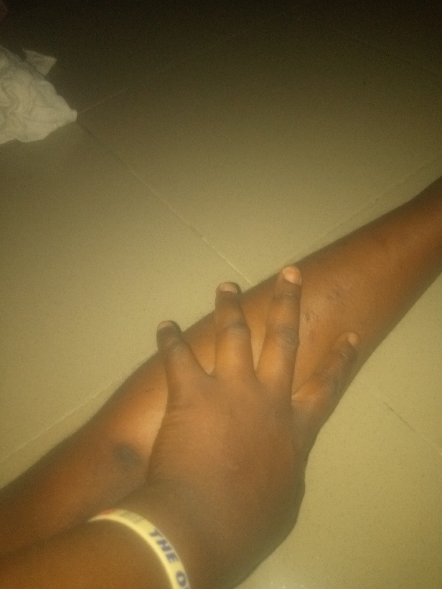 The image depicts a close-up view of a person's forearm and hand resting on a tiled surface. The skin tone is dark. A white bracelet with yellow lettering is visible on the forearm. The tiled floor appears to be a light beige color with visible grout lines. A white cloth is present in the upper left corner of the image, partially out of focus. The lighting is dim, suggesting an indoor environment. There is no information within the image to suggest the location in the city of Jauro Yinu, Nigeria.