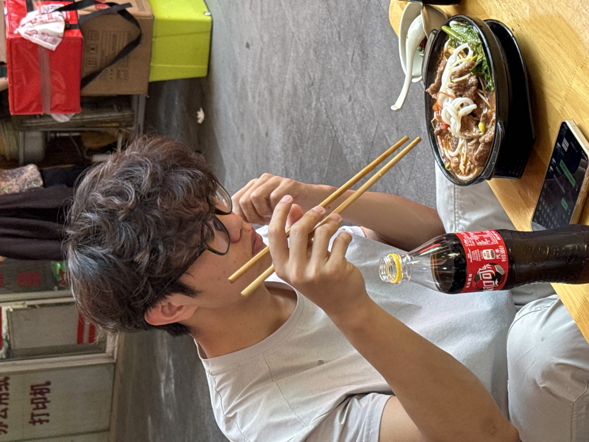 A young man with short, curly brown hair and glasses sits at a wooden table eating noodles with chopsticks in what appears to be an outdoor dining area. He is wearing a light grey t-shirt and white pants. Beside the bowl of noodles is a bottle of Coca-Cola and a smartphone on the table. The location appears to be in Beibei, China based on the Chinese characters visible in the background.