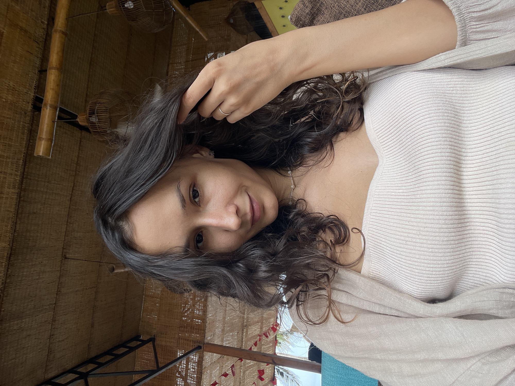 A young woman with long, dark, curly hair is captured in a selfie. She is wearing a light-colored ribbed top and a beige cardigan draped over her shoulders. A thin, pearl necklace is visible around her neck. Her right hand is raised, touching her hair, and she wears a thin ring on her right ring finger. She is looking towards the camera with a slight smile.
The background suggests an outdoor or semi-outdoor setting, possibly a cafe or restaurant with a thatched roof made of woven material. Bamboo structures are visible supporting the roof, and woven lampshades hang from the ceiling. In the background, there is a glimpse of a teal-colored sofa or cushion and some red decorative elements hanging from what appears to be a canopy. The overall ambiance suggests a relaxed, possibly tropical environment. The specific location is identified as Phu Thuy Ward, Vietnam.