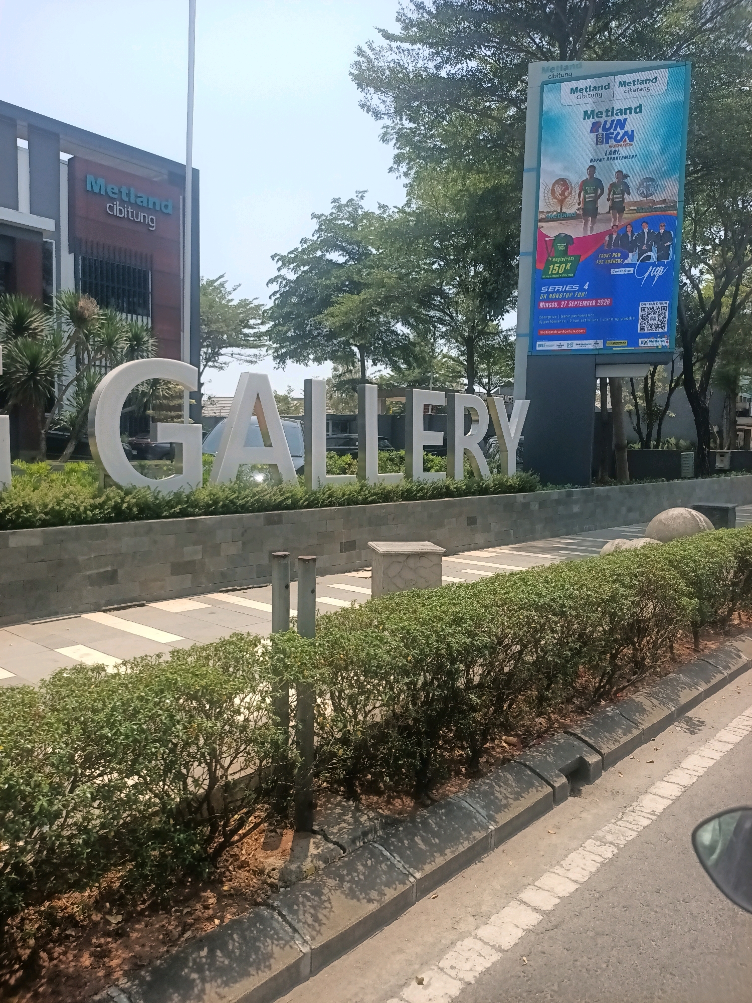 The image shows the "Metland Cibitung" sign, a large three-dimensional white lettering, in front of a modern building. The sign appears to be in the city of Telagaasih, Indonesia. To the right of the sign, a large billboard advertises "Metland Run Fun," featuring images of runners and performers. The area is landscaped with green bushes and trees. The sky is clear and blue. The image is taken from a slightly elevated perspective, likely from a vehicle, as indicated by the presence of a car mirror in the lower right corner.