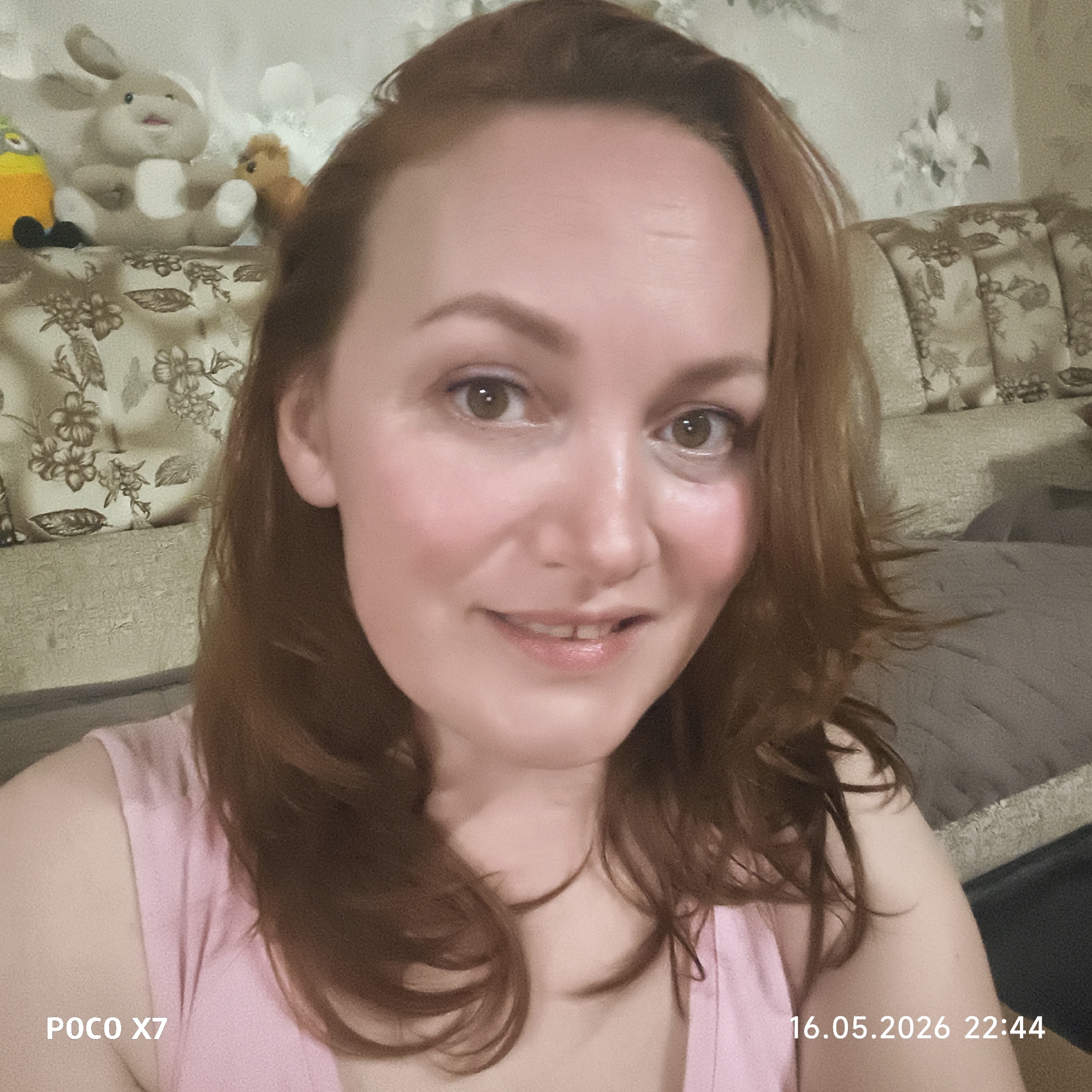 A reddish-haired woman is looking directly at the camera in an indoor setting. She is wearing a light pink top. Behind her is a patterned sofa or wall covering, and a bed or a different part of the sofa is visible. There are stuffed animals on the shelf behind her. "POCO X7" and "16.05.2026 22:44" are visible in the lower corners. While it is not possible to confirm the location in Chaikovsky, Russia, without further context, the scene appears to be an interior setting in someone's home.