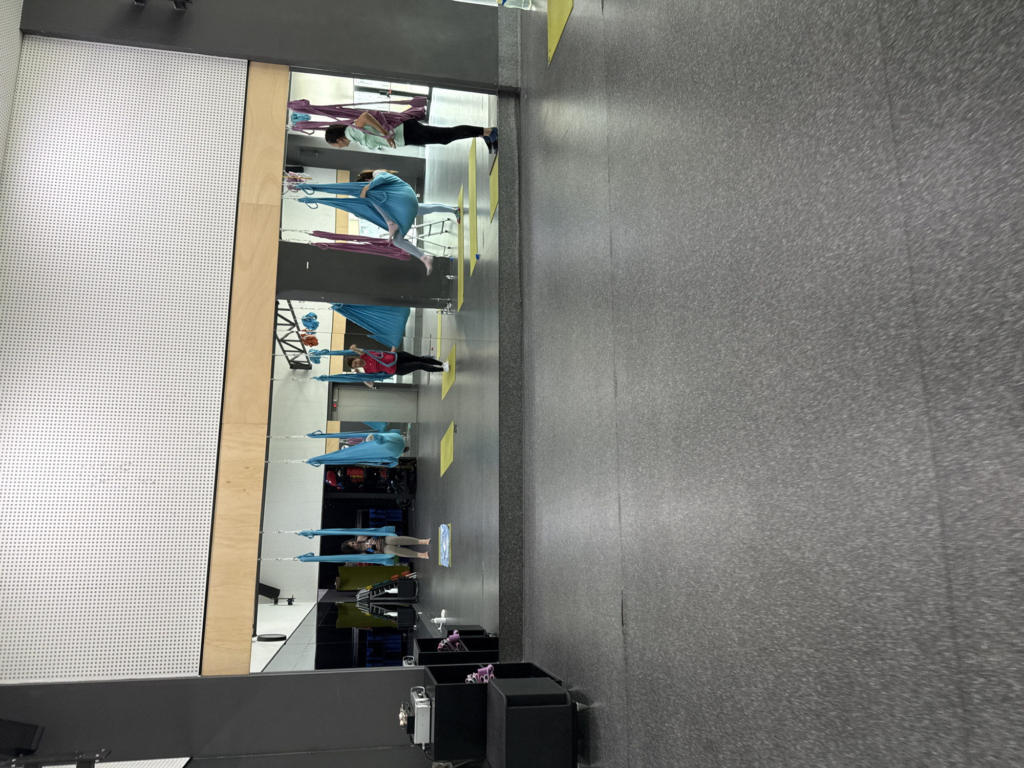 The image shows an indoor aerial yoga studio. In the foreground, a gray, speckled floor dominates the scene. In the mid-ground, a large mirror reflects the activities of the studio. Several individuals are practicing aerial yoga, suspended in colorful hammocks of blue and purple. One person in a red top and black pants stands near a blue hammock. Another person in a light blue top and beige pants is seated in a blue hammock. A third person in a light green top and black pants stands to the right, observing. The studio appears to be well-lit, with a white, perforated ceiling visible in the reflection. To the left of the mirrored area, a black modular furniture unit with shelves is visible, holding various items including what appears to be a projector.