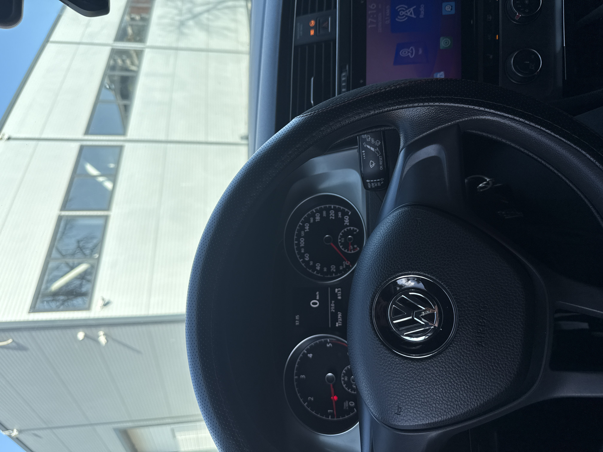 The image shows the interior of a car, specifically the dashboard and steering wheel of a Volkswagen. The steering wheel is black and has the VW logo in the center. The dashboard includes the speedometer, tachometer, and a digital display showing information like time (17:16), odometer (173797), trip meter (813.3), and outside temperature (21.0). The car's multimedia system screen is visible in the top right corner.
Through the car's windshield, a building with rows of windows is visible. The building appears to be a commercial or industrial structure with white siding.
While the image suggests the car is in Varna, Bulgaria, based on the question's premise, the provided visual information does not contain locational clues confirming its exact position. More data, like street signs or distinctive landmarks, would be required to specifically determine the location within Varna.