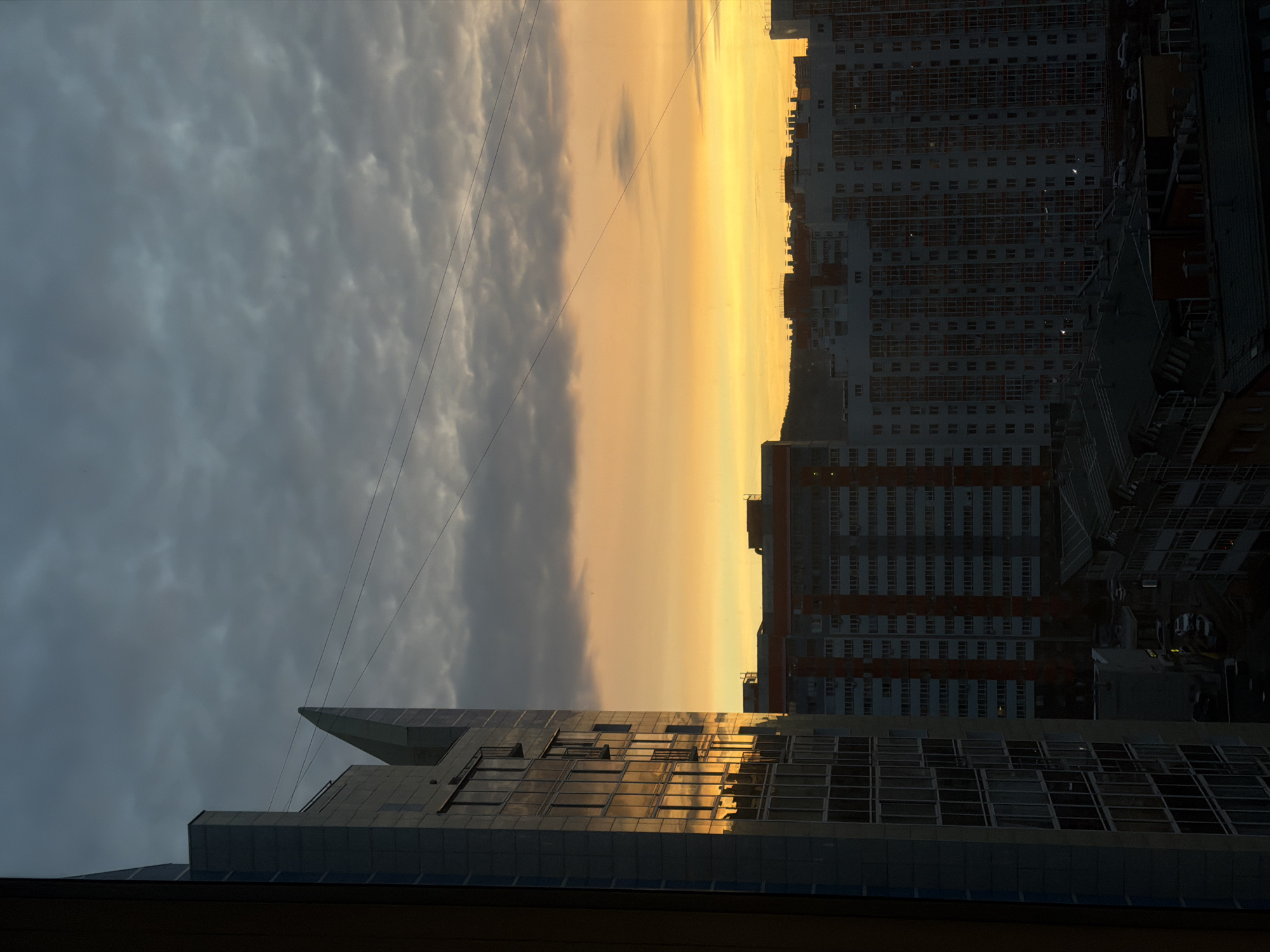 The image captures a cityscape at dawn or dusk in Krasnoyarsk, Russia. The view is from an elevated position, likely an apartment or office building, overlooking a cluster of buildings. The sky transitions from dark, cloudy blues on the left to a bright, yellow-orange band in the center, indicating the presence of the sun near the horizon. To the right, tall residential buildings with numerous windows dominate the skyline. In the foreground, the flat roof of another building, with visible reflections of the sky, is partially visible. Power lines stretch across the sky in the upper-left quadrant.