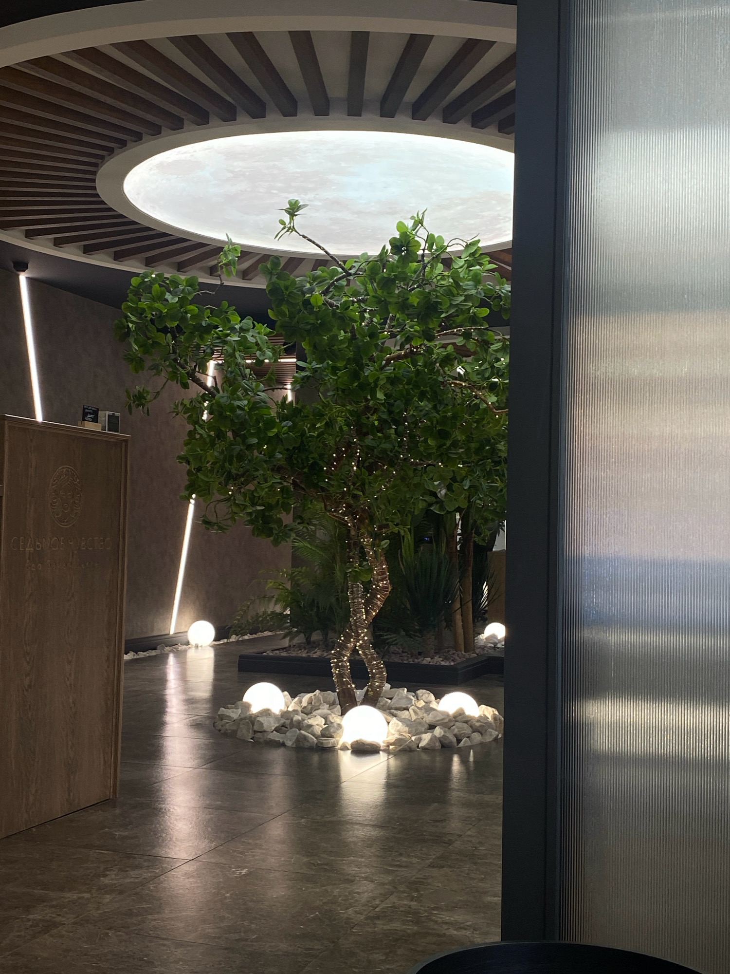 The image shows an indoor space with a prominent artificial tree decorated with fairy lights in the center. The tree is surrounded by white spherical lights and decorative stones. A wooden panel with an emblem and text in Cyrillic, possibly a business name or sign, is visible on the left. The ceiling features a circular light fixture with wooden slats. The floor is tiled and reflects the lighting. On the right, a textured glass partition is present. The overall ambiance is modern and decorative. The location is specified as being in Votkinsk, Russia, though no visual cues in the image directly confirm this. There are no people or animals visible in the scene.