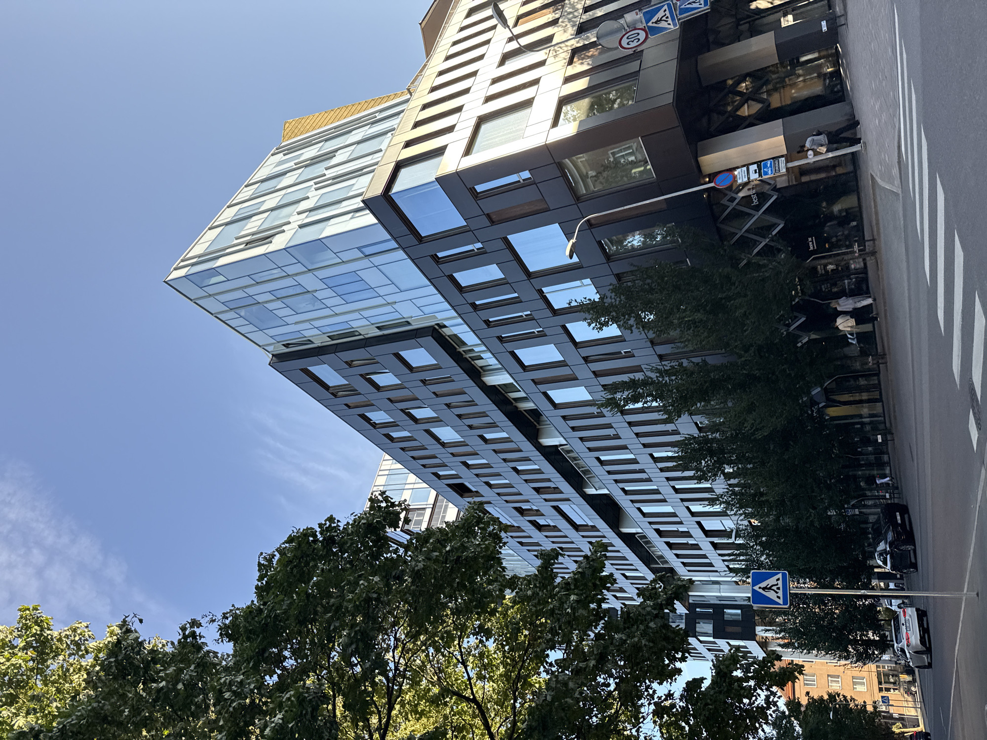 The image shows a modern, multi-story building with a dark facade and glass windows, located in Tallinn, Estonia. The building has a distinctive angled design, with a section clad in glass and another in what appears to be dark metallic panels. Sunlight illuminates the scene from the side, casting shadows. In the foreground, a street with a pedestrian crossing is visible, with a white car and a black car driving on the road. A pedestrian crossing sign and a speed limit sign (30) are present on the sidewalk. Several trees with green foliage line the street in front of the building. A few individuals are visible on the sidewalk and in the vicinity of the building. The sky is blue with some wispy clouds. The overall impression is an urban street scene with contemporary architecture.