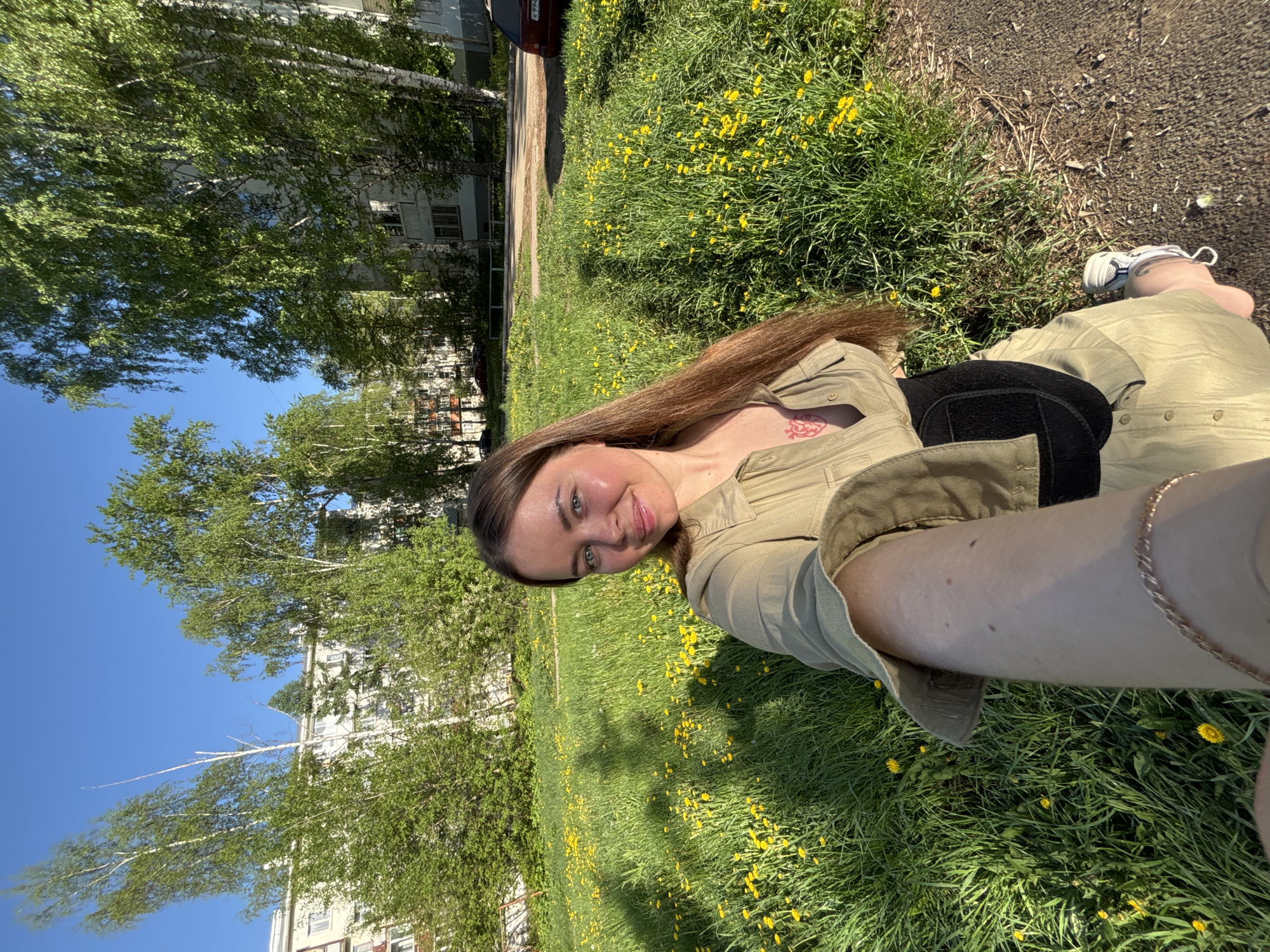 A fair-skinned woman lies in a grassy area with dandelions in Izhevsk, Russia. She is wearing a light green button-down shirt and light green pants, with a black back brace. She has long brown hair and is smiling towards the camera. A tattoo is visible on her upper chest. The background includes green trees and a multi-story building, indicating an urban park or residential area. Part of a dark-colored car is visible on the upper left. A paved area is adjacent to the grass on the right. The sky is blue.