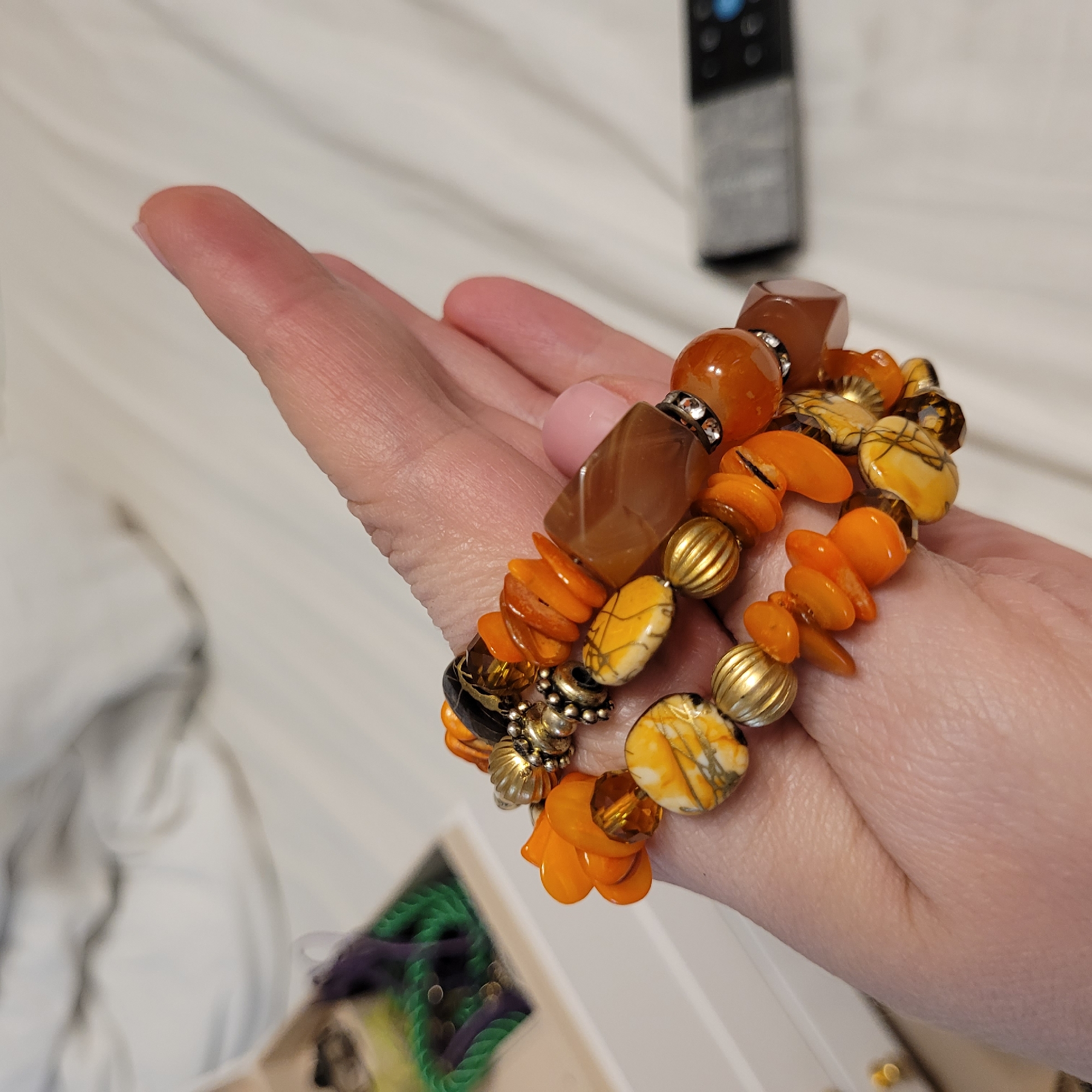 A close-up, high-angle shot features a person's hand holding two stacked, vibrantly colored bracelets against a softly lit, light-colored background. The hand, angled upwards and to the left, has a light pink polish visible on the fingernails.
The two bracelets are adorned with an eclectic mix of beads in warm, autumnal tones, primarily shades of orange, amber, brown, and yellow. Specific bead types include large, smooth, round orange-amber beads, some accented with small, silvery, rhinestone-studded spacer beads. Also present are irregular, faceted amber-brown beads, small flat orange "chip" beads, and round, ribbed, metallic gold-toned beads. Interspersed among these are flat, irregular oval beads with a cream or yellowish base, featuring distinctive dark brown or black crackle patterns. Smaller dark-colored beads and additional metallic spacers contribute to the bracelets' varied textures.
The background consists of a soft, light-colored fabric, likely a white sheet or blanket, exhibiting subtle folds. In the upper right corner, a blurred black and silver remote control rests on this surface. A highly blurred area in the lower left suggests other items, possibly additional jewelry with green and purple components, hinting at an indoor setting such as a bedroom or dressing area. No text is visible in the image. The scene captures a moment of presenting or showcasing the colorful, handcrafted-style jewelry.