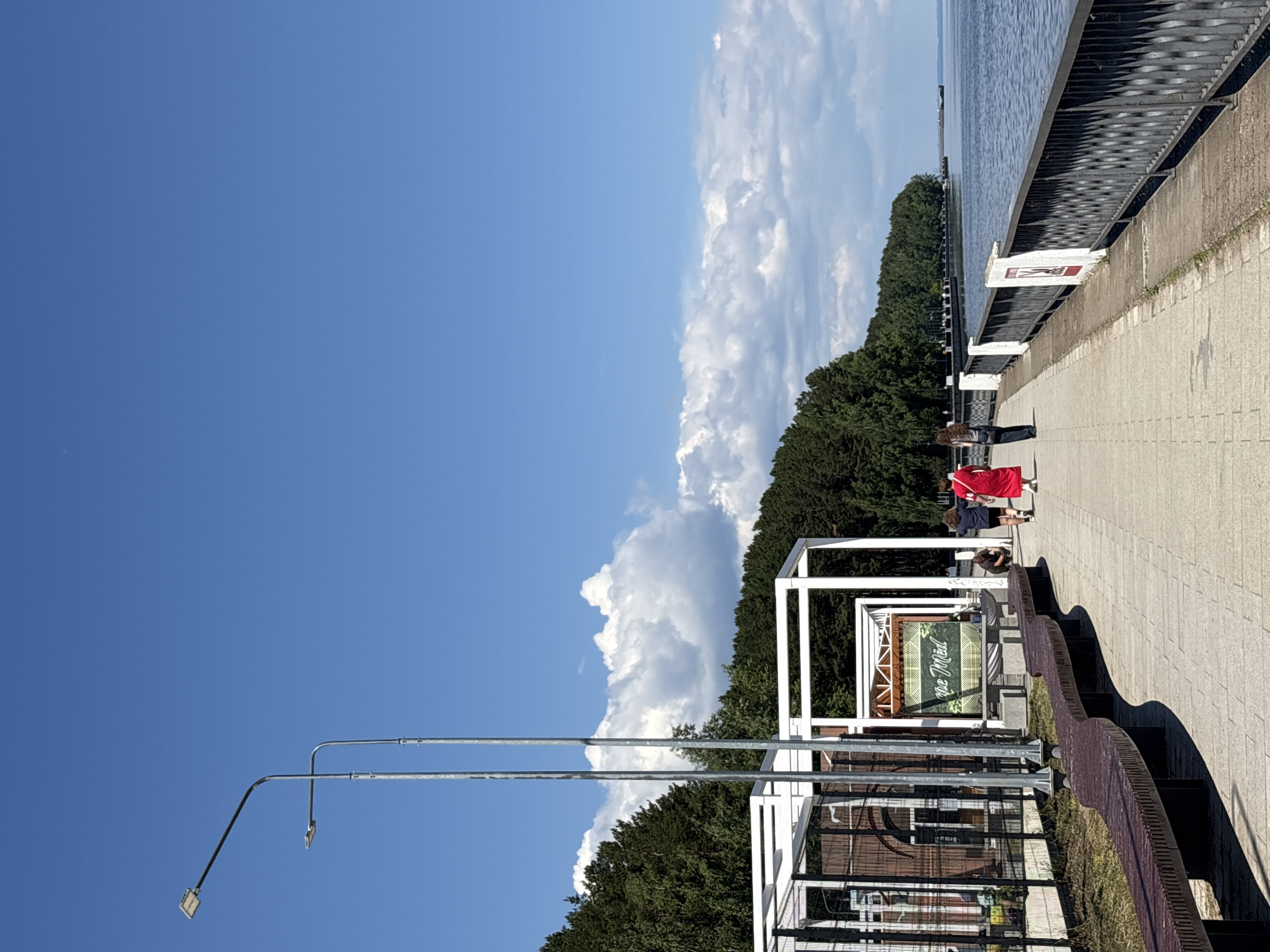The image depicts a sunny day along a waterfront promenade in the city of Chaikovsky, Russia. A paved walkway lines the edge of a large body of water, likely the Kama River, with a dense forest forming a backdrop on the opposite shore. Several white modern structures, possibly pavilions or shelters, are visible on the left side of the promenade, alongside long, dark brown, undulating benches. Two lampposts stand tall on the left.
In the foreground, a long, curved bench stretches along the walkway. A few people are seen walking away from the camera on the promenade. One person in a bright red dress stands out, walking alongside another individual in dark clothing. Further down the promenade, a couple is walking together, and a person is seen crouching near some plants. A few other figures are indistinctly visible further along the path. The water appears calm, with a distant ship visible on the horizon. The sky is a clear blue with scattered white clouds.