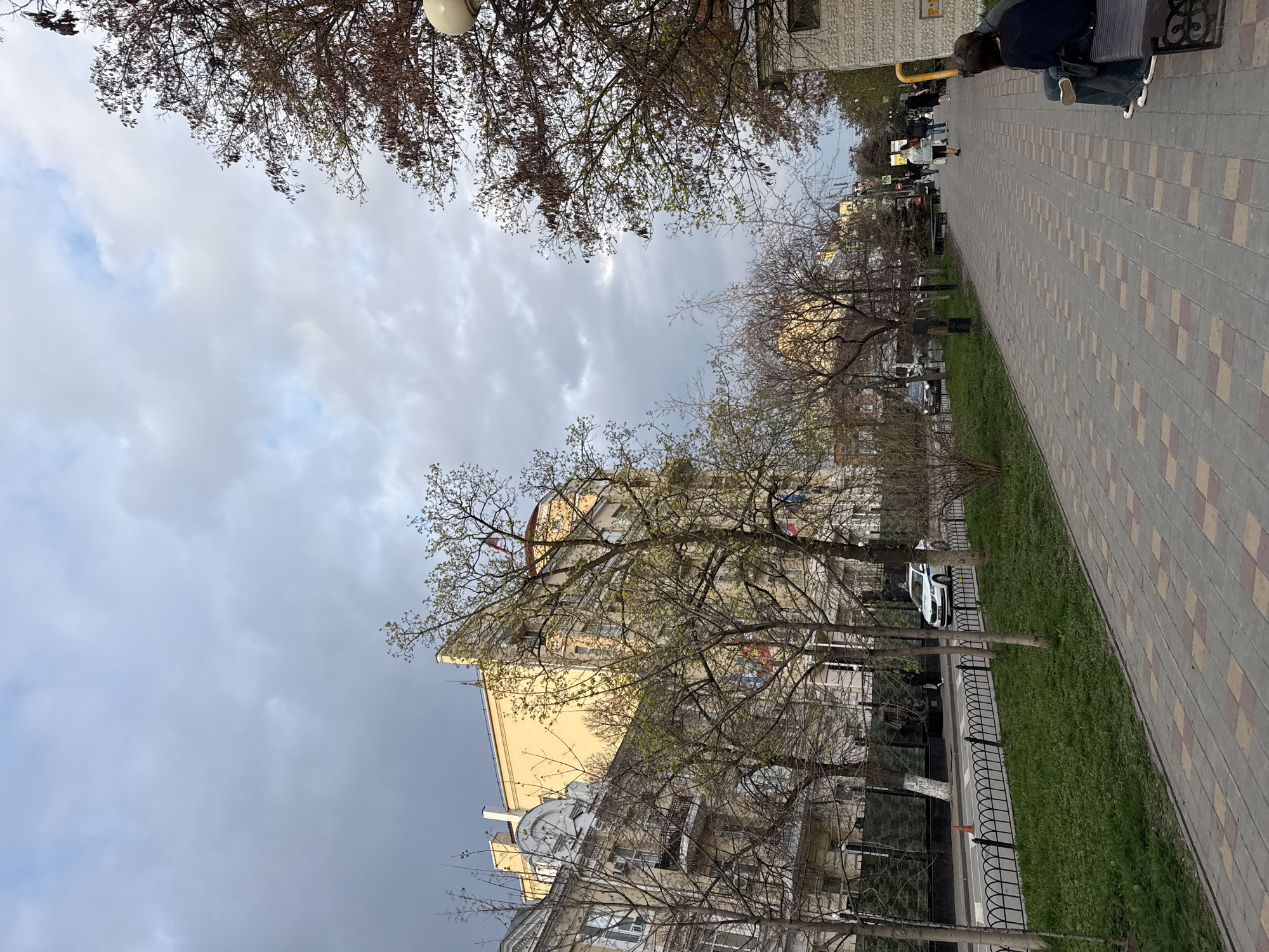 The image captures a street scene in Rostov-on-Don, Russia, under an overcast sky. A paved walkway, patterned with alternating yellow and gray squares, extends into the distance along the right side. A narrow strip of green grass separates the sidewalk from a wrought iron fence that runs parallel to the road. Beyond the fence, trees line the street, some still bare, while others show signs of early spring foliage. Several buildings of varying heights stand on the opposite side of the road, displaying architectural details such as balconies, windows, and decorative elements. A light-colored building with a noticeable peak can be observed amidst the trees. Several people can be seen walking along the walkway. A person sits on a bench, facing away from the camera. Cars are parked along the street. The overall atmosphere suggests a quiet urban scene on a cloudy day.