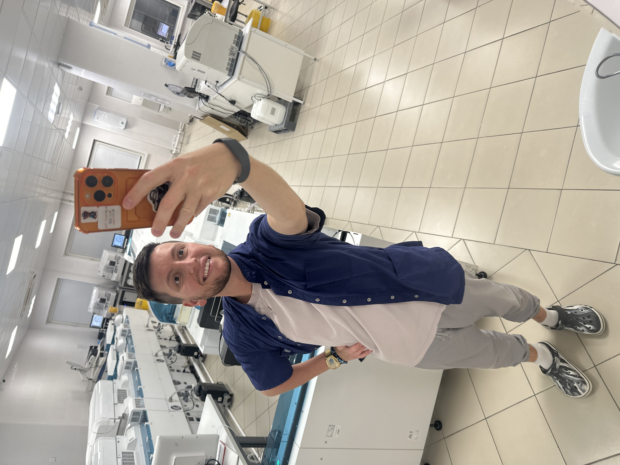 This media file captures a selfie taken by a young man in what appears to be a laboratory or cleanroom setting in Moscow, Russia.
**Subjects:** The primary subject is a young adult male, standing upright and smiling directly at the camera. He has short dark hair and a neatly trimmed beard. His left hand, adorned with a dark smartwatch, holds a smartphone up to take the selfie. His right wrist displays a golden watch and a beaded bracelet.
**Objects:** The man is wearing a light-colored (possibly white or light beige) t-shirt under a dark blue, short-sleeved, collared button-up shirt, worn open. He pairs this with light grey sweatpants, white socks, and distinctive grey and white patterned Crocs with a flame-like design. The smartphone, an iPhone model, is housed in a bright orange case featuring several stickers. The background is dominated by rows of large, white, modern laboratory machines, interconnected by various cables and featuring multiple computer monitors. The floor is covered with large, light-colored square tiles. A white sink or basin is partially visible on the right edge. Fluorescent light fixtures illuminate the white walls and ceiling.
**Activity or event taking place:** The man is engaged in the act of taking a selfie, evident from his arm outstretched holding the phone and his direct gaze into the lens.
**Notable details:** The environment is brightly lit, pristine, and highly organized, characteristic of a professional scientific, medical, or industrial laboratory. The sophisticated equipment suggests automated analysis or processing. The man's smiling expression conveys a sense of happiness or contentment. The time of day and weather are not discernible due to the indoor setting.
**Visible text:** Text on the phone case stickers includes "WTF?!" and a cartoon character. The brand "CROCS" is visible on his footwear. Text on the golden watch is too small to discern.