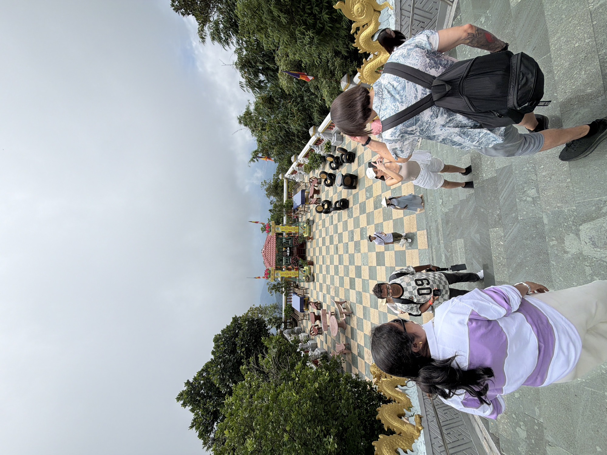 The image shows a group of people walking up a wide stone staircase that leads to a courtyard area. The courtyard has a checkered floor with yellow and green tiles. In the background, there is a colorful temple structure with red roofs and yellow pillars. Surrounding the courtyard are lush green trees and some distant mountains visible under a cloudy sky. Golden dragon sculptures are visible along the sides of the staircase.
The people in the foreground are walking up the stairs. The person closest to the camera is wearing a white and purple horizontally striped shirt and light-colored pants. To their left and slightly behind is another person from the back, wearing a blue and white patterned shirt and gray shorts, with a black backpack. To the left of the person in the striped shirt, a man with glasses and a checkered shirt with the number "09" is visible. Further up the stairs, a woman in white shorts and a light-colored top is taking a photo with her phone. Another person in a light blue dress and a conical hat is also visible.
The overall scene suggests a tourist attraction, possibly a temple or cultural site, in a mountainous or elevated area. Given the visual cues like the temple architecture, dragon motifs, and the surrounding vegetation, this location is highly likely to be the Linh Ung Pagoda on Son Tra Peninsula in Da Nang, Vietnam. The pagoda is known for its large Buddha statue and its panoramic views of the city and the East Sea, and it features elaborate architecture and dragon carvings consistent with the image. The presence of tourists also aligns with this being a popular visitor site.