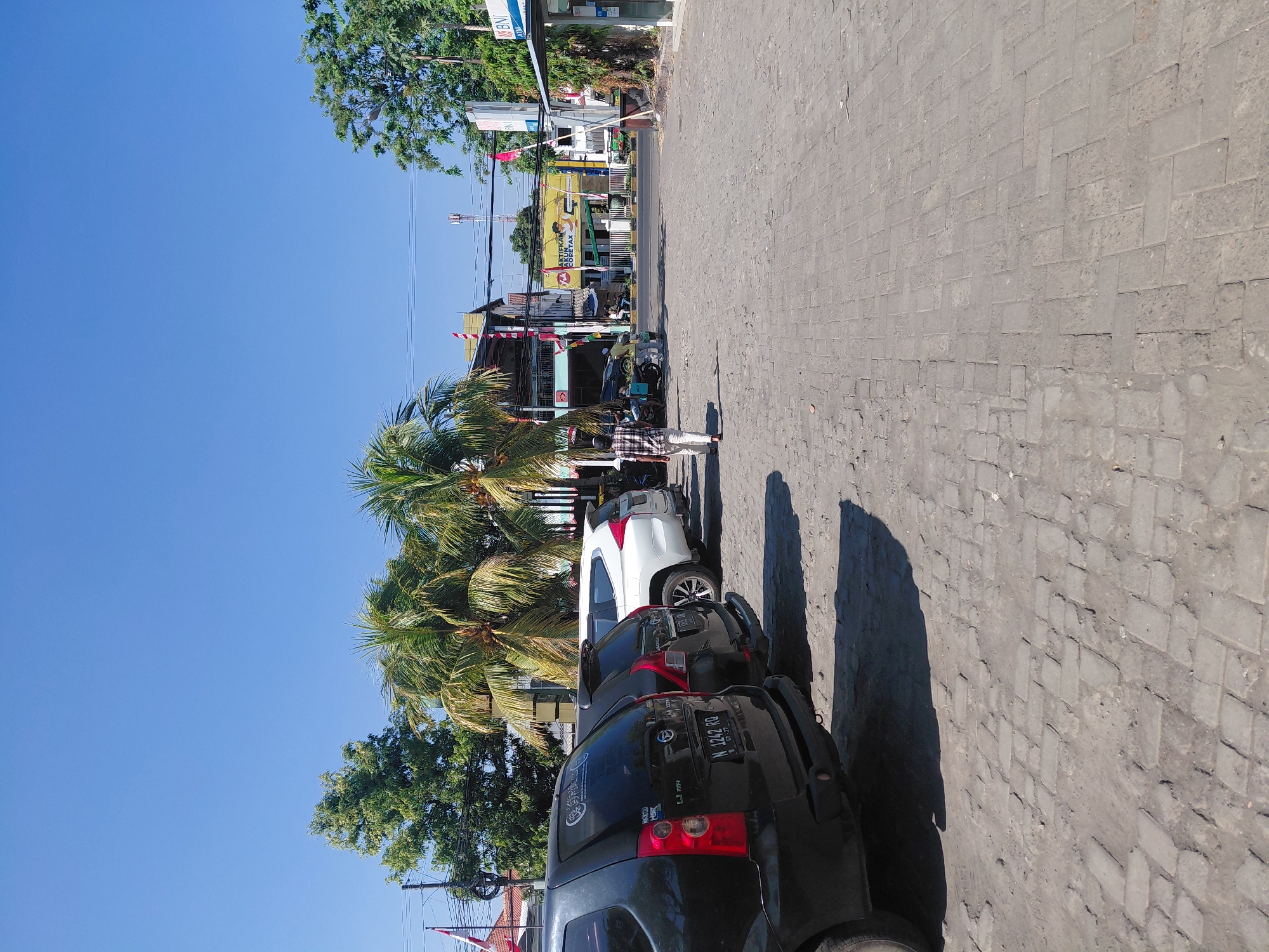 The image depicts a sunny daytime scene in the Semampir area of Indonesia. In the foreground, a paved lot made of interlocking bricks is visible, with shadows cast by parked vehicles. To the left, the rear ends of two black cars are prominent, with the license plate "N 1242 RO" on the closer one. Further back, a white SUV is parked. A person in a checkered shirt and white pants is walking away from the camera towards the right.
Behind the parked cars, a line of buildings and businesses is visible. One building is yellow with red and white signage that includes the text "AKTIFKAN E-FIN COPETAX". Another building has the "BNI" logo and "ATM" displayed. Palm trees are scattered throughout the scene, adding to the tropical ambiance. The sky above is clear and blue. The overall impression is of a commercial area with everyday activities taking place.