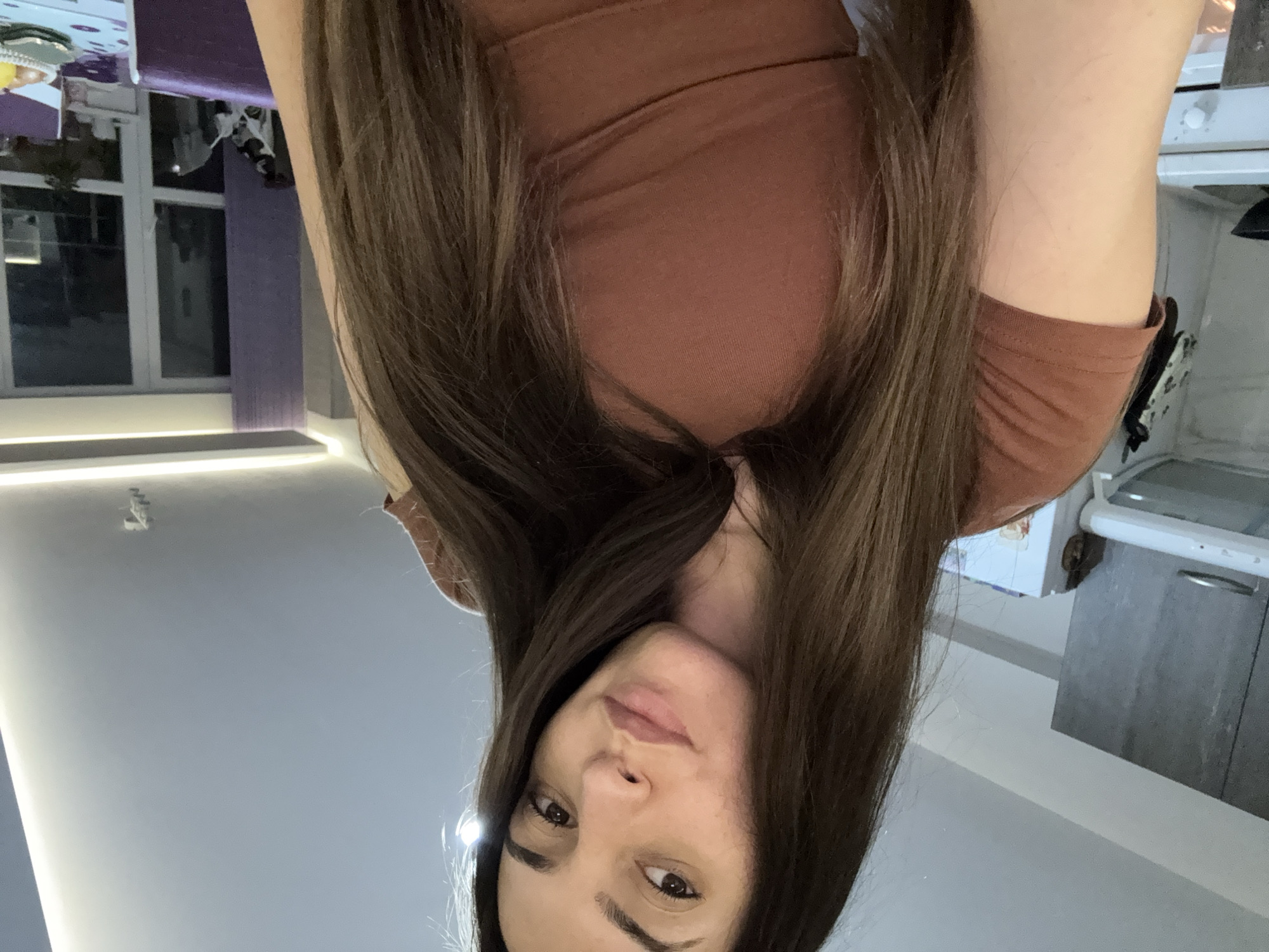 The image shows a woman with long brown hair, wearing a brown top, taking a selfie indoors. The background features a room with a window on the left, and a kitchen area on the right with a white countertop and gray cabinets. It's difficult to determine the specific location within Plovdiv, Bulgaria, without additional contextual cues.