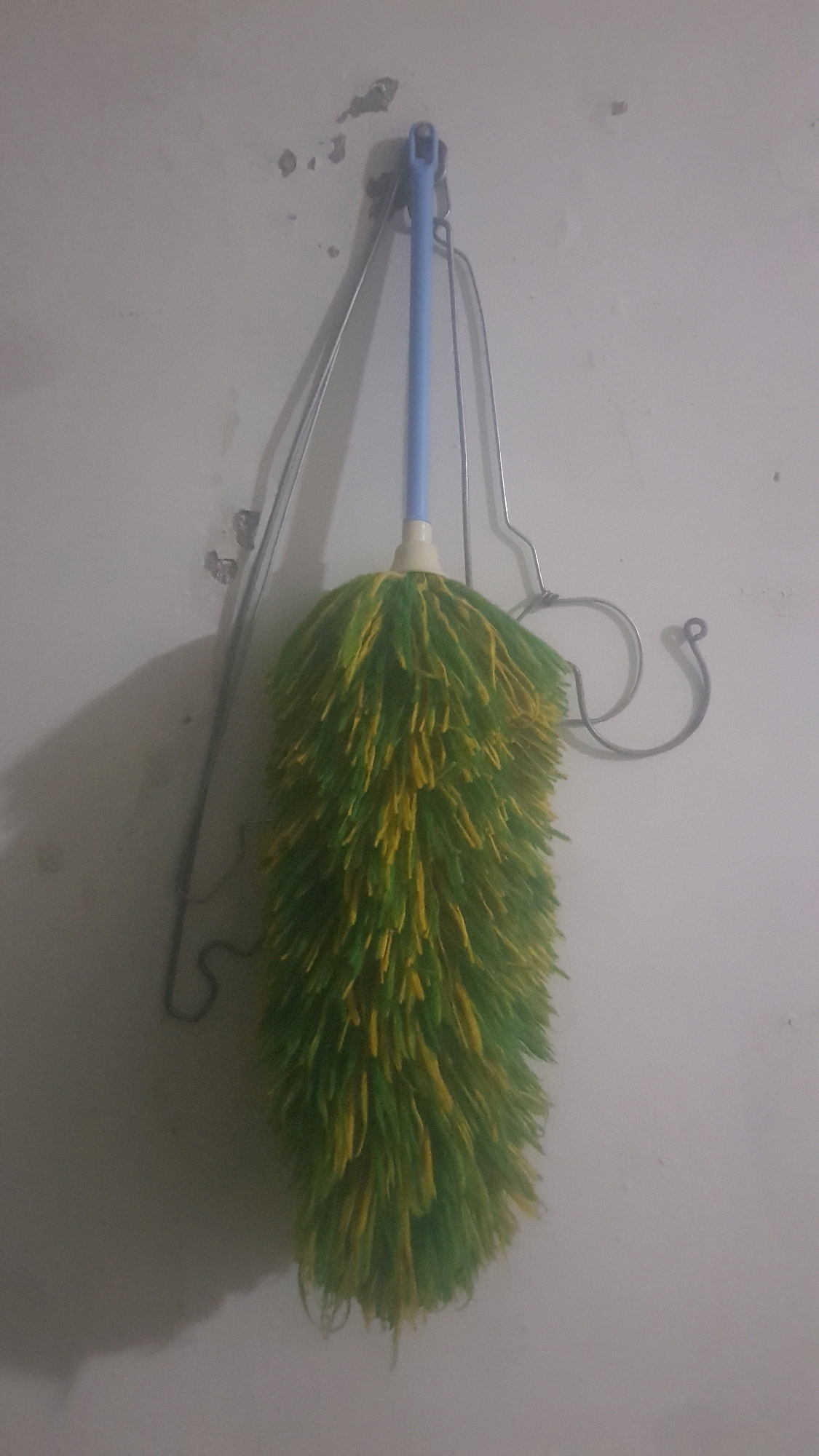 A fluffy, green and yellow duster hangs from a hook attached to a textured, off-white wall. The duster is suspended by its light blue handle, which is connected to a metal hook system. The hook system itself appears to be made of bent metal rods, one of which is looped into a circular shape. The wall shows signs of wear, with some small patches of discoloration and chipped paint. A shadow of the duster is cast on the wall to the left of the object. The image appears to be taken indoors, possibly in a home or storage area. The location is specified as Bakalan, Indonesia.