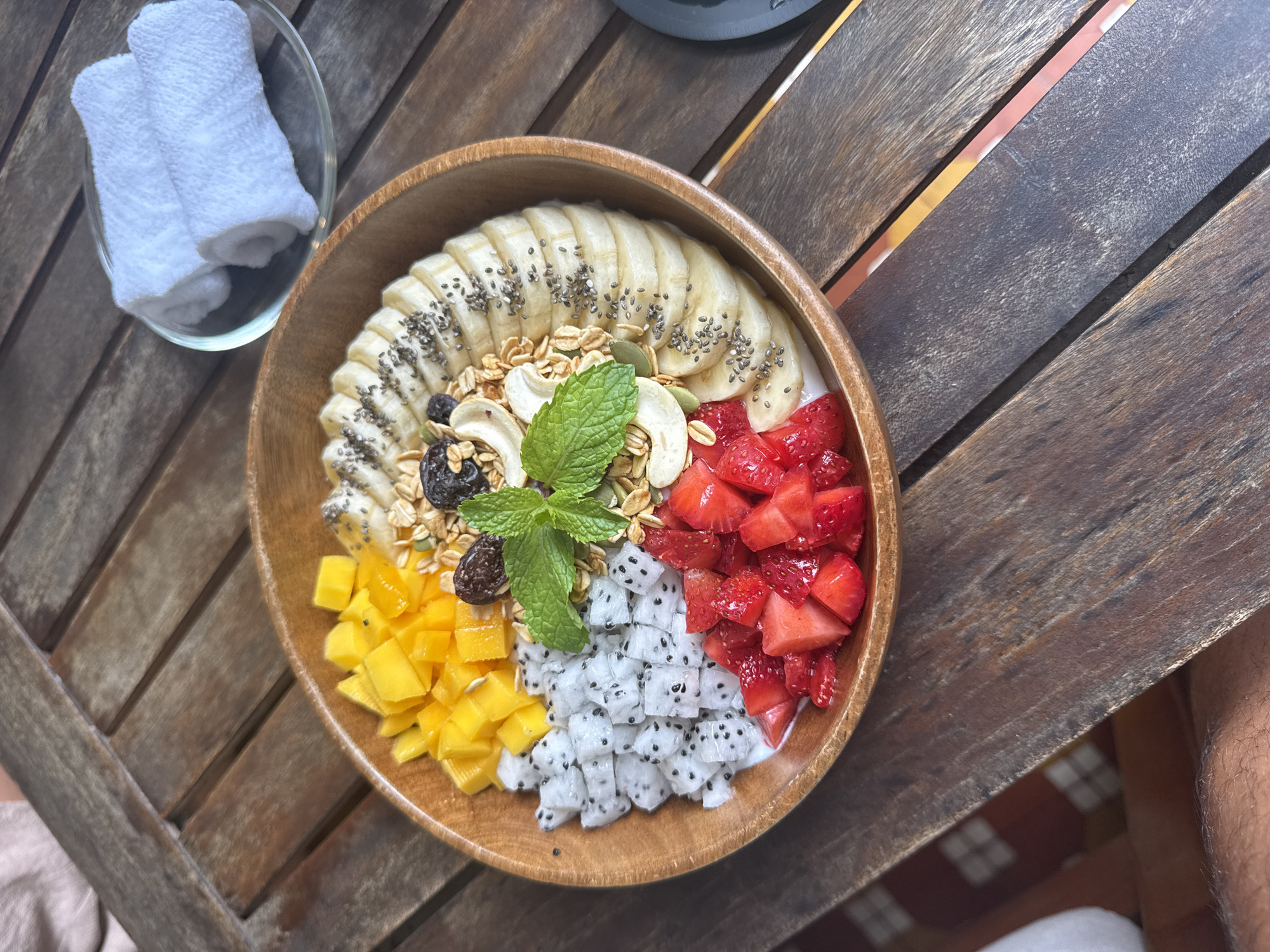 A wooden bowl of fruit and granola sits on a brown wooden table with gaps in the slats. Inside the bowl, clockwise from the top, are sliced bananas topped with chia seeds, granola with cashews, sliced strawberries, cubed dragon fruit, and cubed mango. Three sprigs of mint are in the center. A small glass bowl with two rolled-up white cloths is next to the fruit bowl. Part of a water bottle and a human leg are visible in the upper right and lower right corners, respectively. The scene may be in Đà Nẵng, Vietnam.