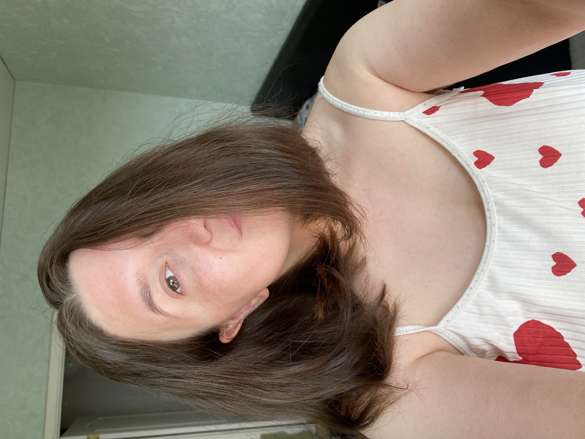 A medium shot portrait of a young woman with shoulder-length brown hair, partially covering her face, as she looks towards the camera. She is wearing a white camisole adorned with red hearts. The background appears to be an indoor setting with a textured green wall and a glimpse of a doorway and a dark object, possibly a headboard. The lighting suggests it is daytime. The location is identified as Chaikovsky, Russia.
