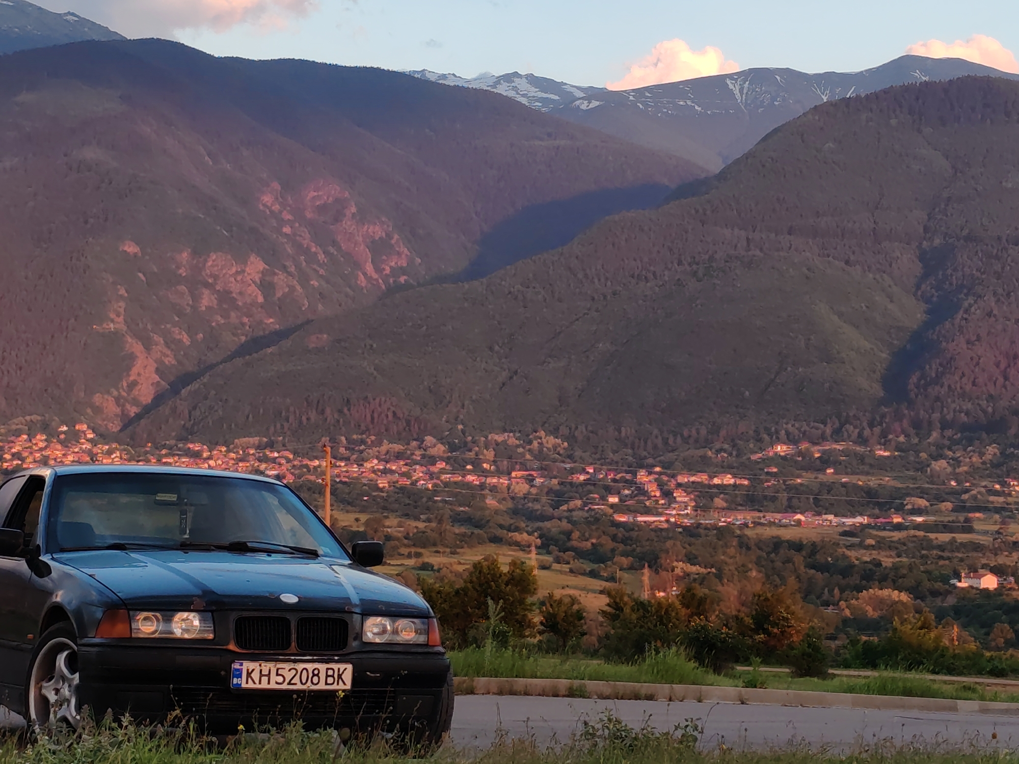 A black BMW E36 sedan is parked in the foreground of a scenic view. The car's license plate reads "KH5208 BK" and indicates it is from Bulgaria. Behind the car is a valley with a town or village, and in the distance, tall mountains are visible, some with snow-covered peaks. The image is taken during late afternoon or early evening light, creating warm tones on the mountains and the village. The location is likely overlooking Dupnitsa, Bulgaria, based on the landscape and license plate. The car is positioned on a paved road or parking area with vegetation near the roadside.