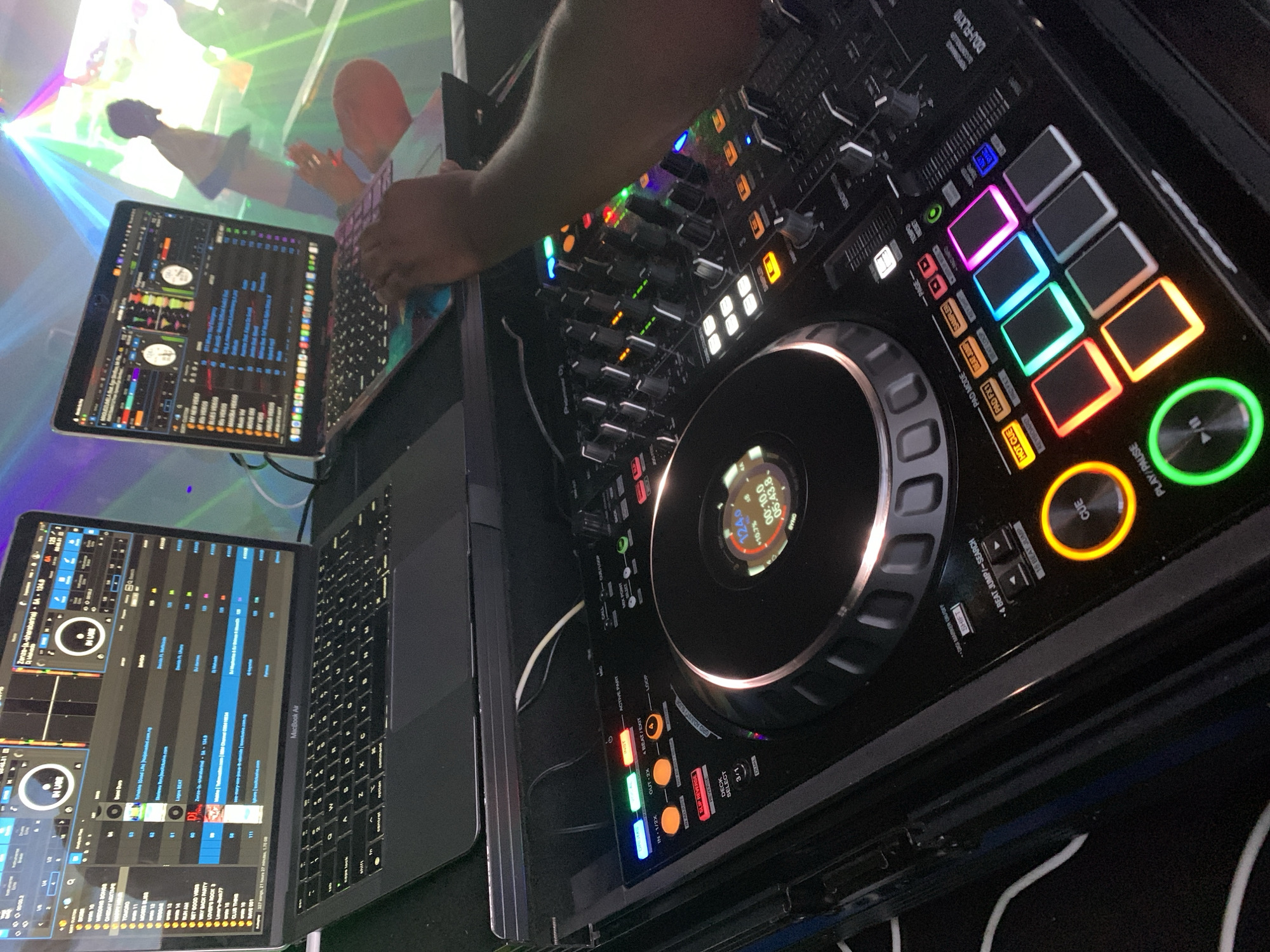 The image depicts a DJ setup in a dimly lit environment, likely a club or event space. Two MacBook Air laptops are positioned side-by-side, displaying DJ software with waveform visualizations and track lists. In the foreground, a Pioneer DJ controller is prominently featured, with its various buttons, knobs, and illuminated pads. A person's hands are visible, interacting with the DJ controller and the laptop. The background shows a blurred scene with colorful lights, suggesting a party or performance atmosphere. The presence of a DJ setup, laptops, and the overall ambiance indicate a music event in progress. The location is stated to be Howayi, Nigeria, suggesting an urban or semi-urban setting within the country.