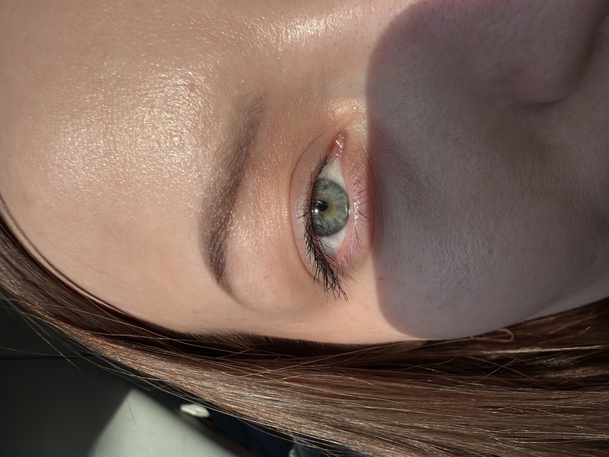 A close-up, slightly rotated view focuses on a person's face, featuring their eye, eyebrow, and surrounding skin. The eye is light-colored with visible black eyeliner applied to the upper lid. The eyebrow is dark. The skin on the forehead and cheek appears smooth. The person has light brown hair visible at the bottom. The image does not provide any contextual information about Minino, Russia.