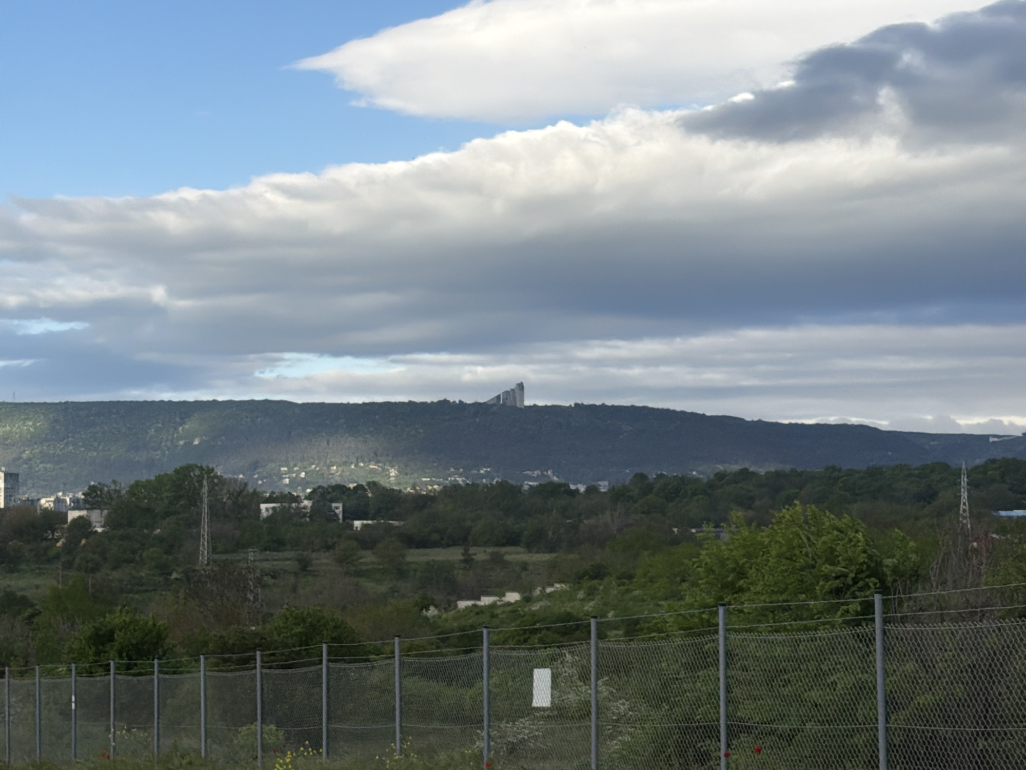 The image depicts a landscape with a chain-link fence in the foreground. Beyond the fence is a field and trees. In the midground, there are rolling hills covered in trees with some buildings visible. The monument Founders of the Bulgarian State is situated atop the highest hill. The sky is partly cloudy. The location is in the city of Shumen, Bulgaria.