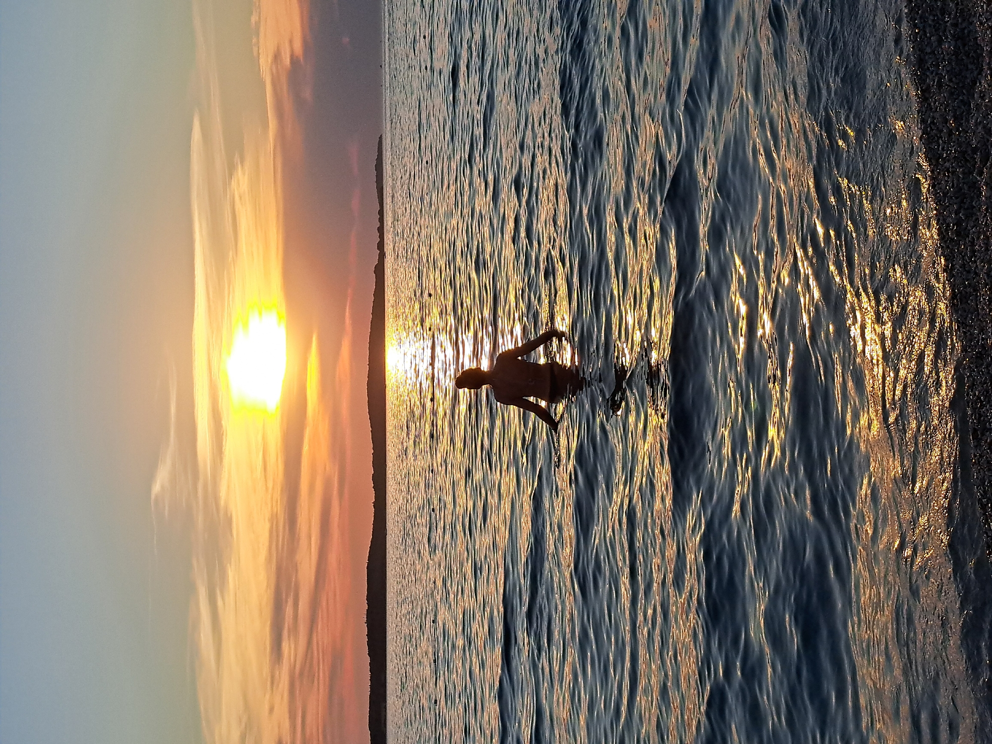 This is a vertical, eye-level, medium shot of a person wading into the water during sunset. The scene depicts a tranquil beach at dusk, with the sun casting a warm, golden glow across the sky and water. The person, silhouetted against the vibrant sunset, appears to be enjoying a refreshing dip. The water is calm, with gentle ripples reflecting the fiery hues of the setting sun. The beach itself is covered in dark pebbles. The overall mood is one of peace and serenity, capturing the beauty of nature at its finest. The time of day is clearly sunset due to the dramatic colors in the sky and the low angle of the sun. The weather appears to be clear and pleasant. No text is visible in the image.