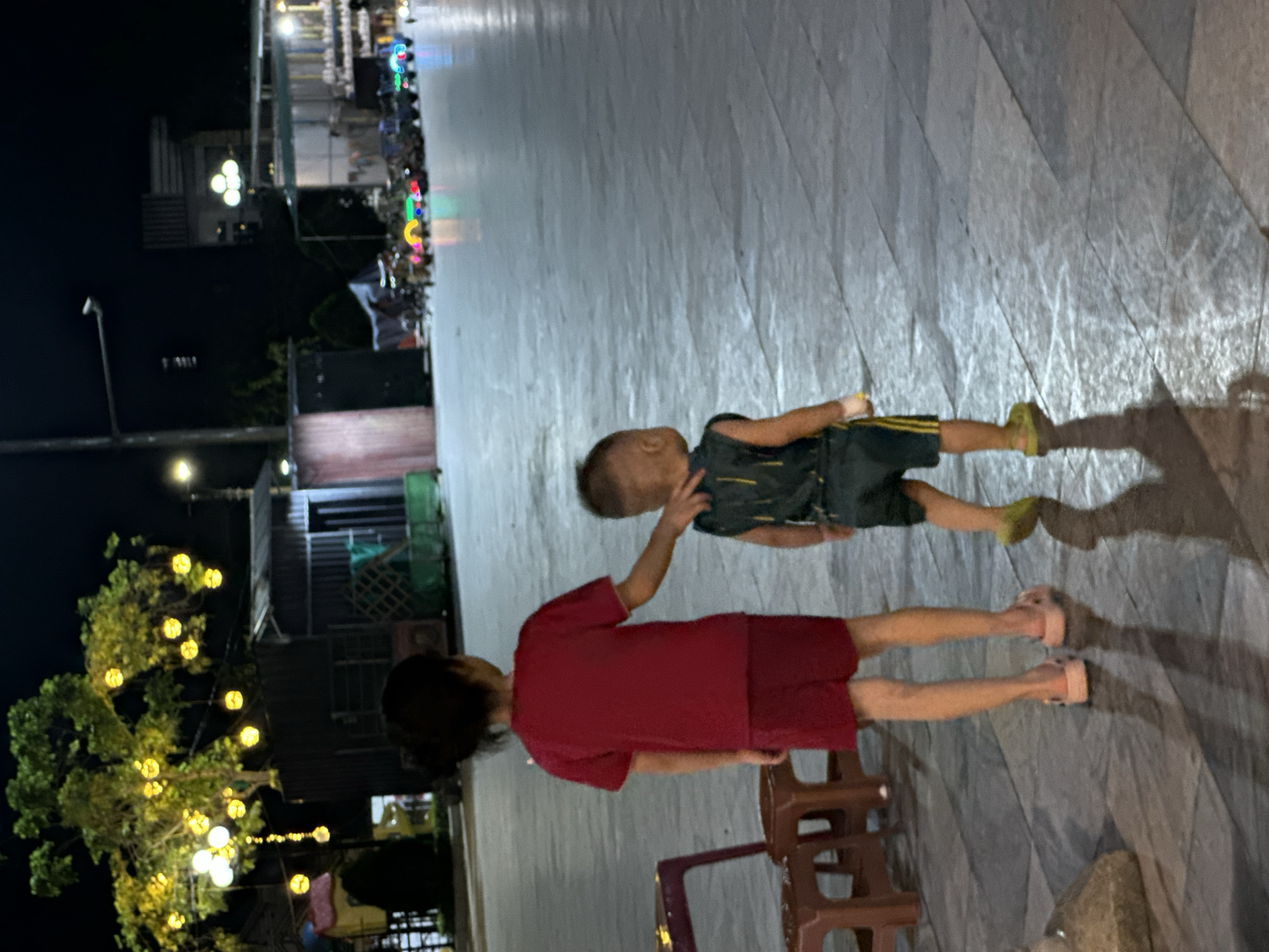 The image captures an outdoor night scene in Phố Ràng, Vietnam, featuring two individuals on a paved surface. In the foreground, an adult or older child, seen from behind, wears a red short-sleeved shirt and red shorts, with light pink sandals on their feet. Their right hand is gently placed on the left shoulder of a younger child standing in front of them. This younger child wears a dark-colored top and shorts, with yellow-green shoes, and is looking slightly to their right, away from the camera. Shadows of both individuals are cast on the ground, suggesting a light source.
The setting appears to be a public square or a wide paved area, featuring light-colored, patterned tiles. Partially visible near the adult's feet are two brown plastic stools. The background is dark but illuminated by various artificial lights. On the left side, a tree is heavily adorned with numerous warm yellow string lights. Further into the background, streetlights and additional buildings are visible, some with illuminated windows or balconies. In the distance, a line of colorful lights suggests a lively street or market area. The overall atmosphere is that of a casual evening gathering or stroll. No visible text is present in the image.