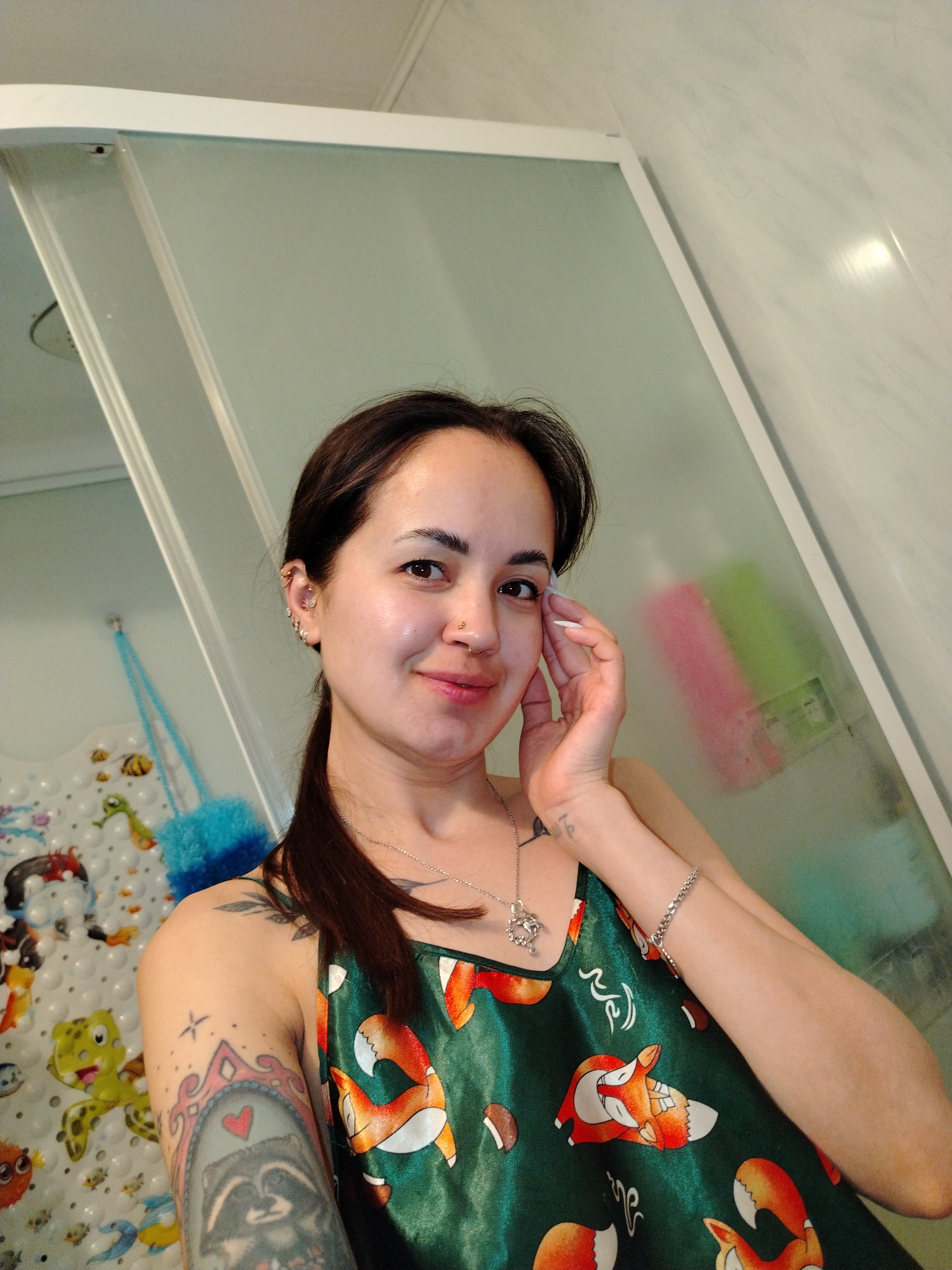 A woman is taking a selfie in what appears to be a bathroom. The woman has medium-brown hair pulled back into a low ponytail, a fair complexion, and multiple piercings in her left ear, as well as a nose ring. She is wearing a green satin tank top with a pattern of orange foxes and mushrooms, a silver necklace, and a silver bracelet. She has tattoos on her left arm and upper chest. Her right hand is raised and touching the side of her face. The background includes a glass shower door with pink and green items visible inside, a bath mat with cartoon characters on it, and a blue mesh sponge. The bathroom appears to be tiled with white marble. The location in the city of Noviy, Russia is not discernible from the image.