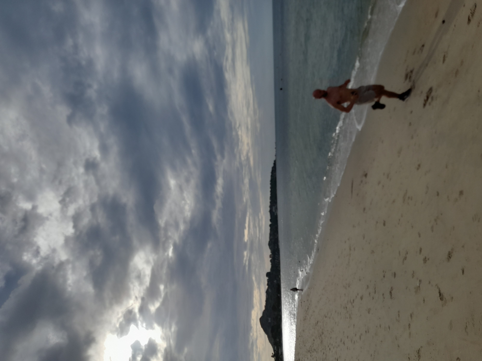 An outdoor, slightly blurred, full shot shows a man running on a sandy beach. He is facing left, with his right leg forward, in mid-stride. He is wearing light-colored shorts and black shoes. His arms are bent at the elbows. Behind him, the sea is calm with small waves lapping at the shore. In the distance, a tree-covered hill or mountain rises from the horizon. The sky is filled with dramatic, dark clouds with patches of sunlight breaking through. Another person is visible in the far distance on the left, walking towards the sea. The sand is light brown and has numerous footprints. The image appears to have been taken during daylight, likely in the late afternoon or early morning due to the lighting conditions.