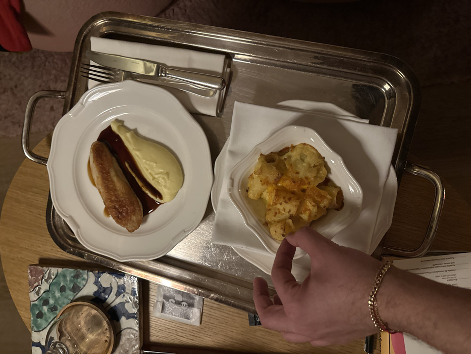 An overhead view captures a prepared meal served on a polished silver rectangular tray within an indoor setting. A male's left hand, wearing a gold-colored chain bracelet, is poised over a side dish, indicating engagement with the meal.
The tray holds a white, scalloped-edge plate featuring a single piece of cooked poultry, likely a chicken breast, presented diagonally over a portion of smooth, light-yellow purée (consistent with mashed potatoes). A rich, dark brown sauce is drizzled across the poultry and purée. To the right of this main dish, a silver fork and knife with classic designs are neatly arranged on a folded white napkin. Below, a smaller, white scalloped-edge bowl contains a gratin-style potato dish characterized by a browned, possibly cheesy, top, resting on another folded white napkin.
The entire arrangement is situated on a light-colored wooden table. To the upper left of the tray, a square ceramic tile with a blue and green ornamental pattern supports a small, clear glass with a wooden base. A small white paper sachet is visible adjacent to the tray. In the lower-left corner, a partially visible document, identified as a menu or bill by discernible text including "Restaurant," "Paris," and "Hôtel," confirms the location as a hotel restaurant in Paris, France. The background beyond the immediate table area is softly blurred, indicating an ambiently lit environment.