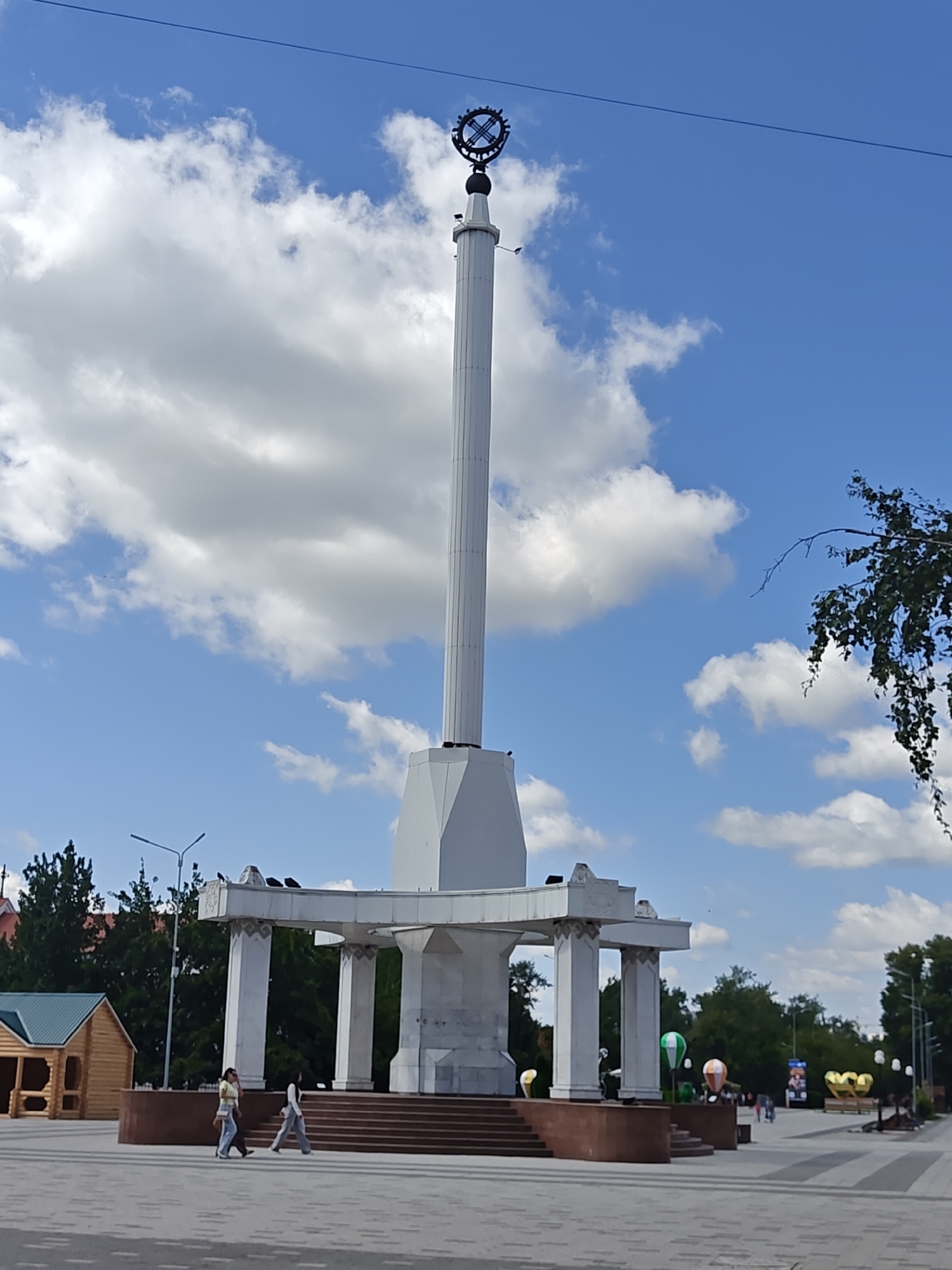 The image shows a large, white monument in Petropavl, Kazakhstan, under a bright blue sky with scattered white clouds. The monument is a tall, cylindrical obelisk topped with a circular, metallic emblem. At the base of the obelisk is a multi-tiered, white, open structure supported by columns, with stairs leading up to it.
In the foreground, a paved plaza is visible, with two young women walking across it. To the left, a small, wooden, gazebo-like structure with a green roof is present. Further back, beyond the monument and trees, are buildings, streetlights, and what appear to be decorative balloons, including some shaped like hot air balloons and gold hearts. The overall scene depicts a public space in the city on a clear day.