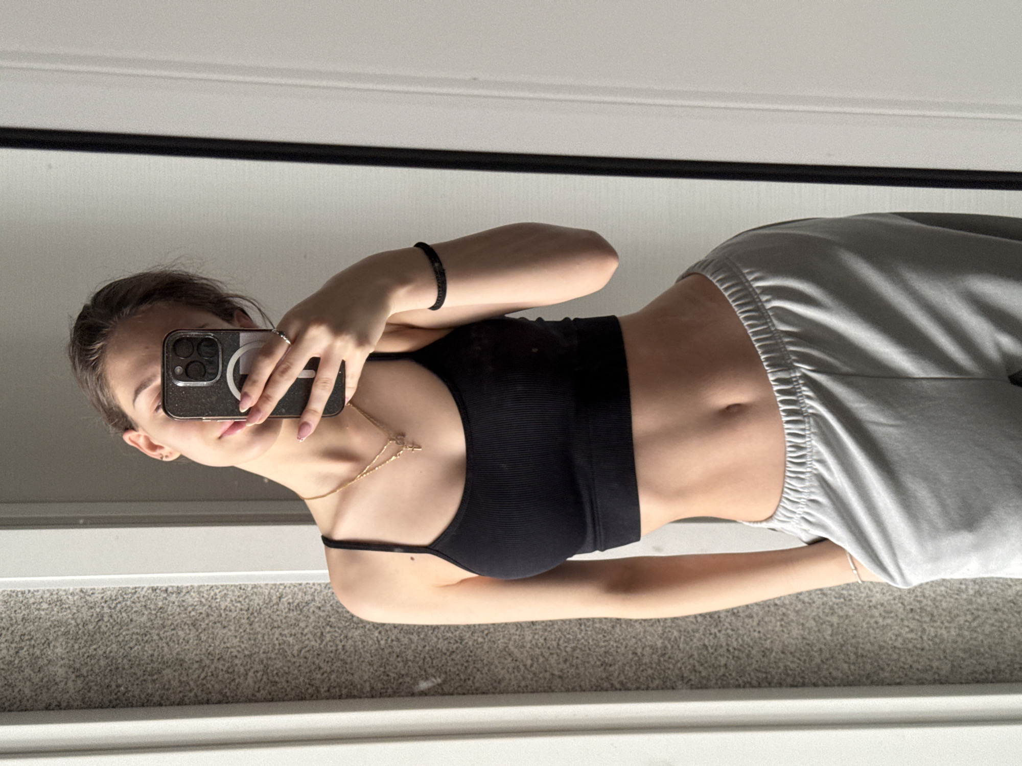 A woman is taking a mirror selfie in what appears to be a room with a light-colored carpet. She is wearing a black crop top and gray sweatpants. She holds a smartphone up to her face, obscuring her features. A gold necklace is visible around her neck, and a black bracelet is on her wrist. The location in Sofia, Bulgaria is not determinable from the image.
