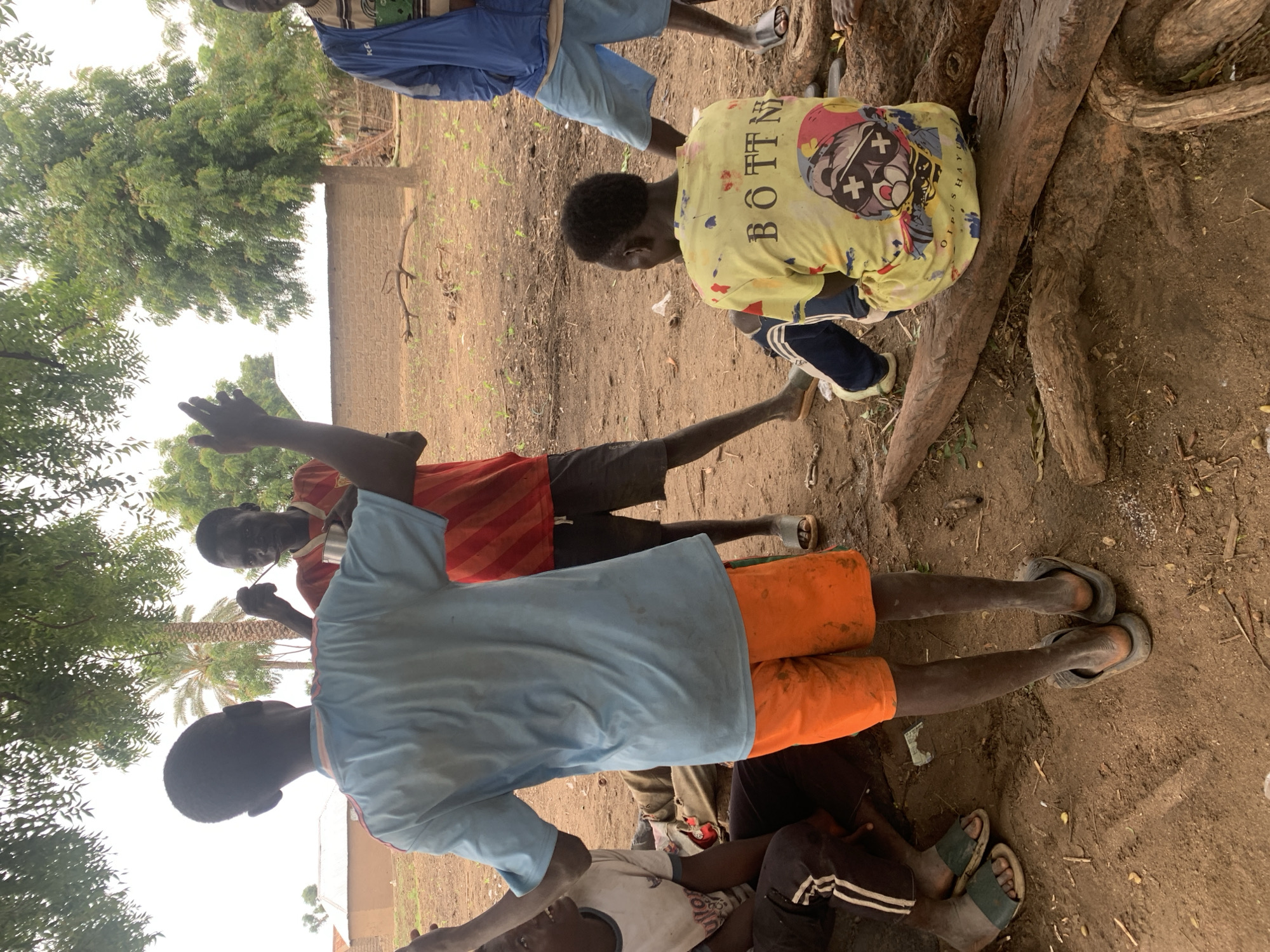 The image depicts a group of young people gathered outdoors in what appears to be a rural setting in Dull A, Nigeria. The ground is dusty earth, with some dry vegetation and tree roots visible. In the background, there is a brick wall and trees with lush green foliage, suggesting it might be a warm day with some shade from the trees.
Several individuals are visible, mostly wearing casual clothing. One person in the foreground is wearing a light blue t-shirt and bright orange shorts, with dark sandals. To their left, another person is wearing a red and white striped t-shirt and dark shorts. Further back, a person in a yellow t-shirt with a graphic print is kneeling near a large tree trunk. This yellow t-shirt has visible text that reads "BÖTTN" and some other words that are less clear. Another individual in the background is wearing blue shorts.
The people appear to be interacting, though the specific activity is not clear. Some are looking towards each other, while others are looking downwards. The overall mood seems relaxed and informal. The lighting suggests it is daytime, possibly mid-morning or afternoon, with diffused light filtering through the trees. The scene conveys a sense of community and everyday life in a Nigerian village.